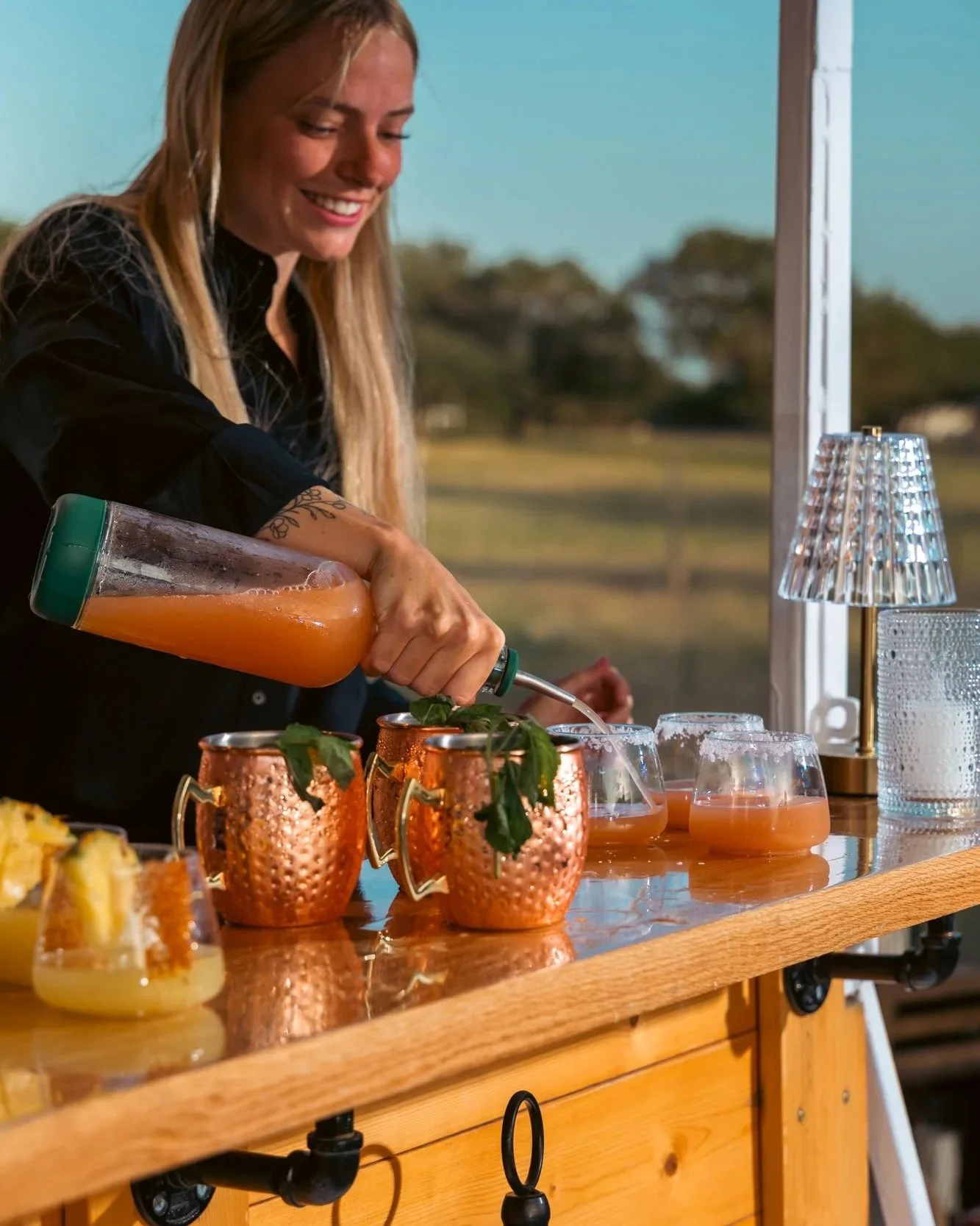 A woman with long blonde hair pouring orange juice into glasses at a wooden bar or table, with decorative copper mugs and various drinks around her, set in a sunny indoor space with a large window and a scenic outdoor background.
