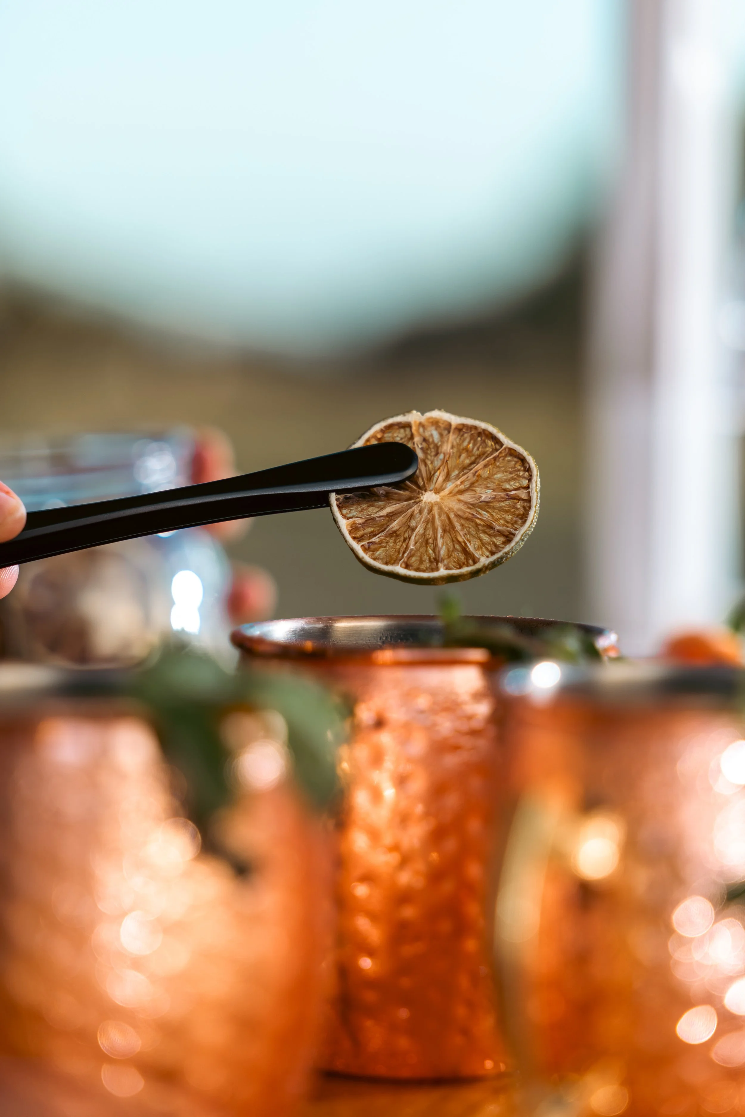 Close-up of a dried lemon slice held with black tongs above a copper mug, with a blurred background of other copper mugs and light bokeh.