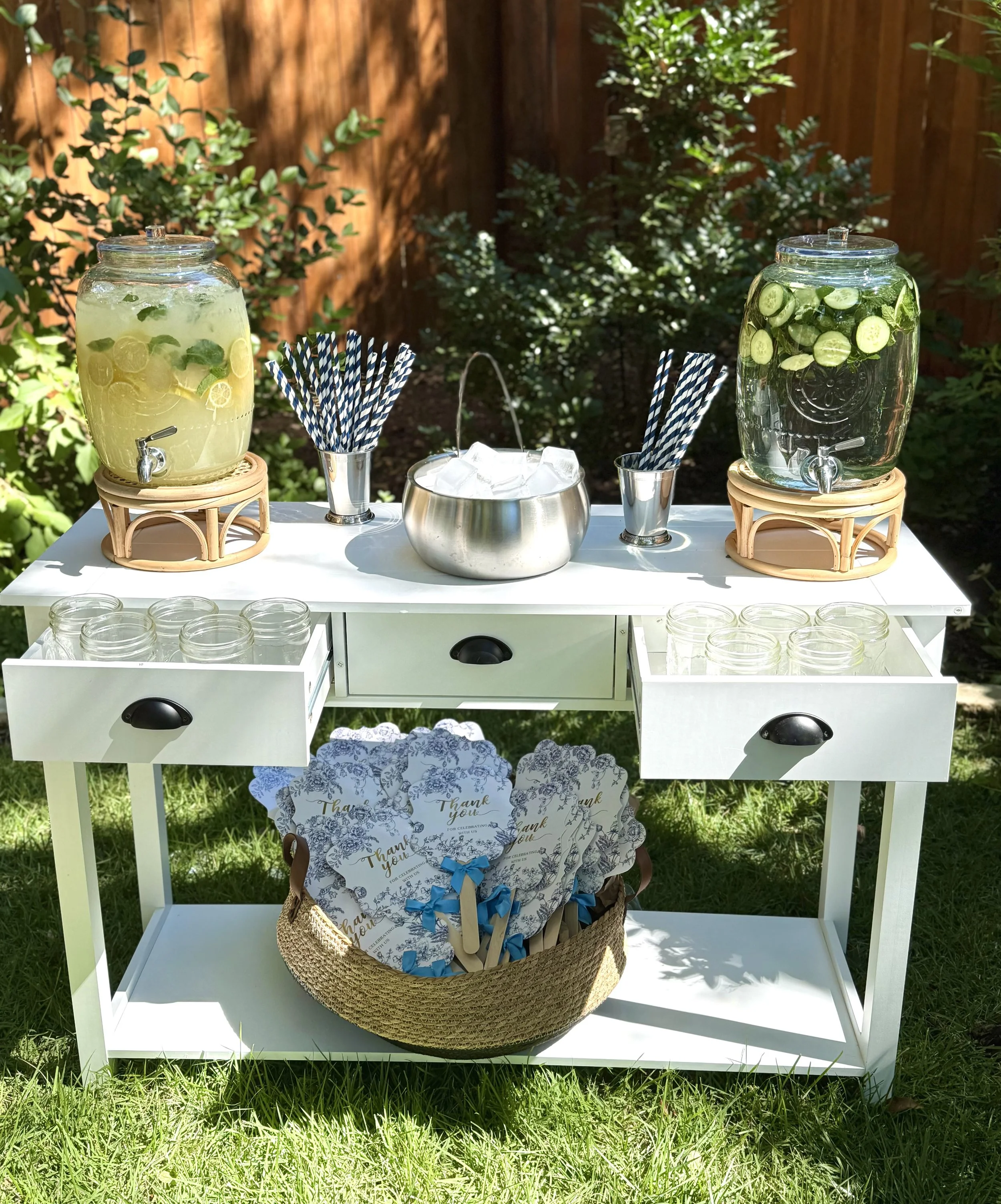Outdoor drink station with two large glass dispensers filled with lemon and cucumber-infused water, paper straws, glass jars, and thank you cards in a woven basket.