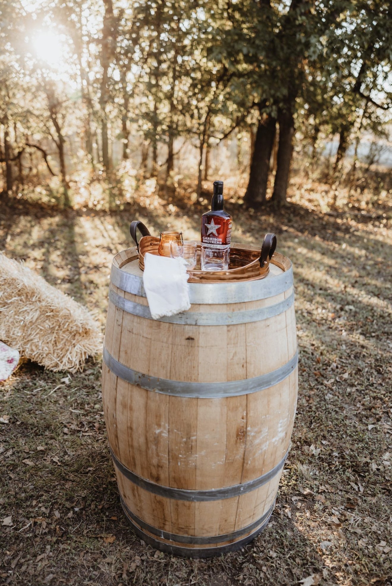 A wine barrel used as a table outdoors in a wooded area during sunset, with a bottle of Garrison Brothers whiskey, two glasses, a napkin, and a small ceramic pitcher on top.