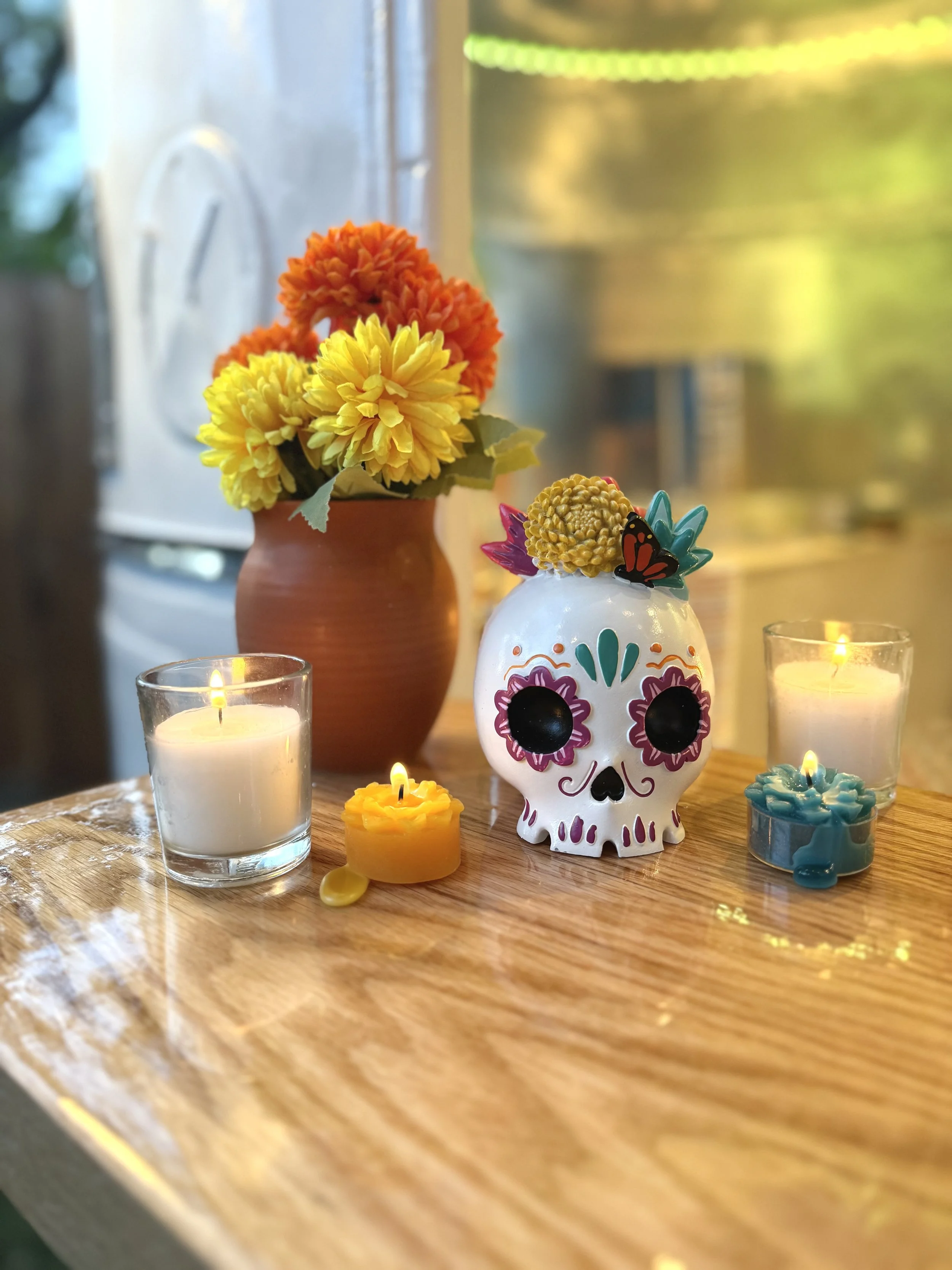 Decorative arrangement on wooden table includes a white skull with painted floral designs, surrounded by lit candles and a vase of orange and yellow flowers in the background.