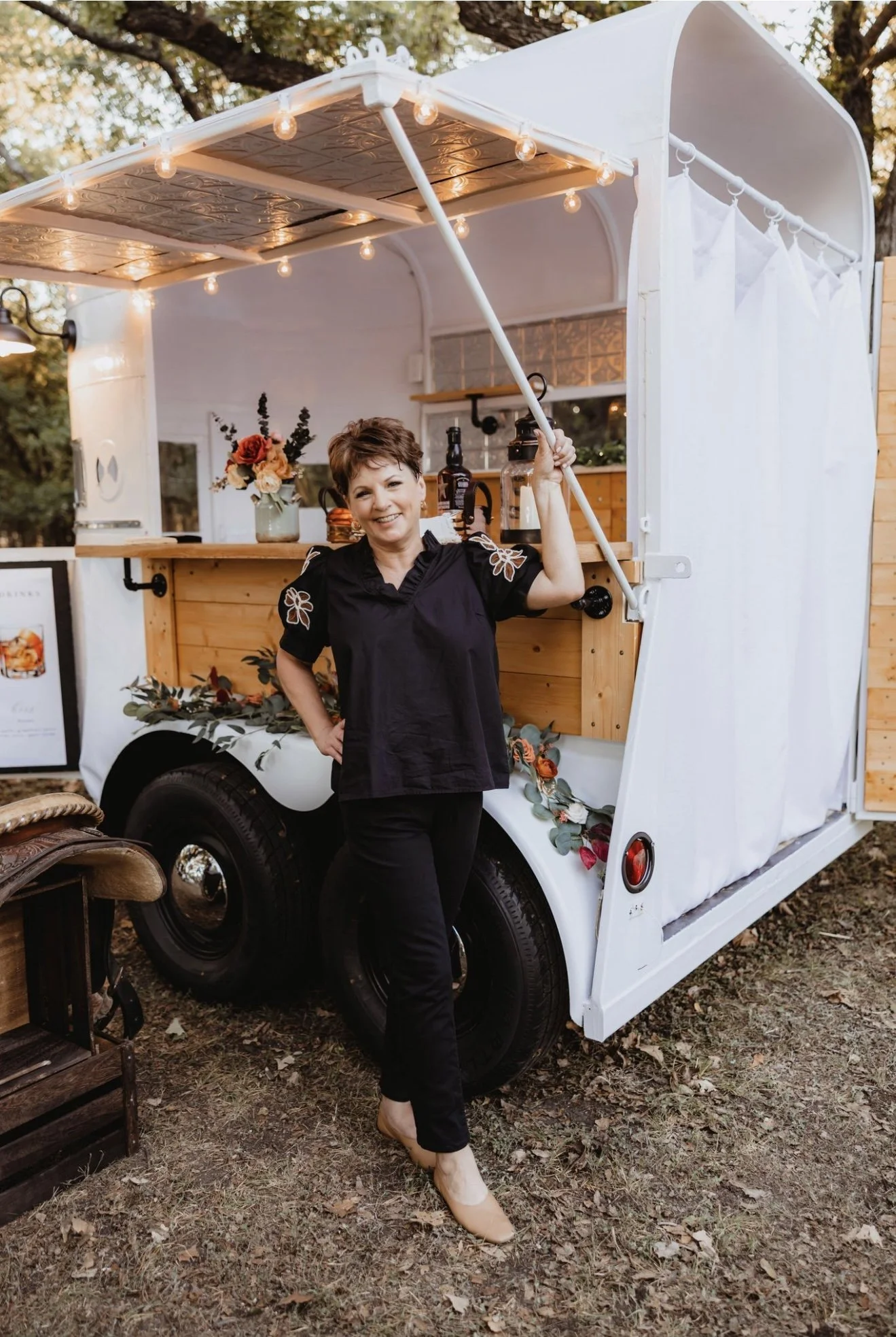 A smiling woman in black standing in front of a mobile bar trailer with string lights, flowers, and bottles, outdoors on grass.