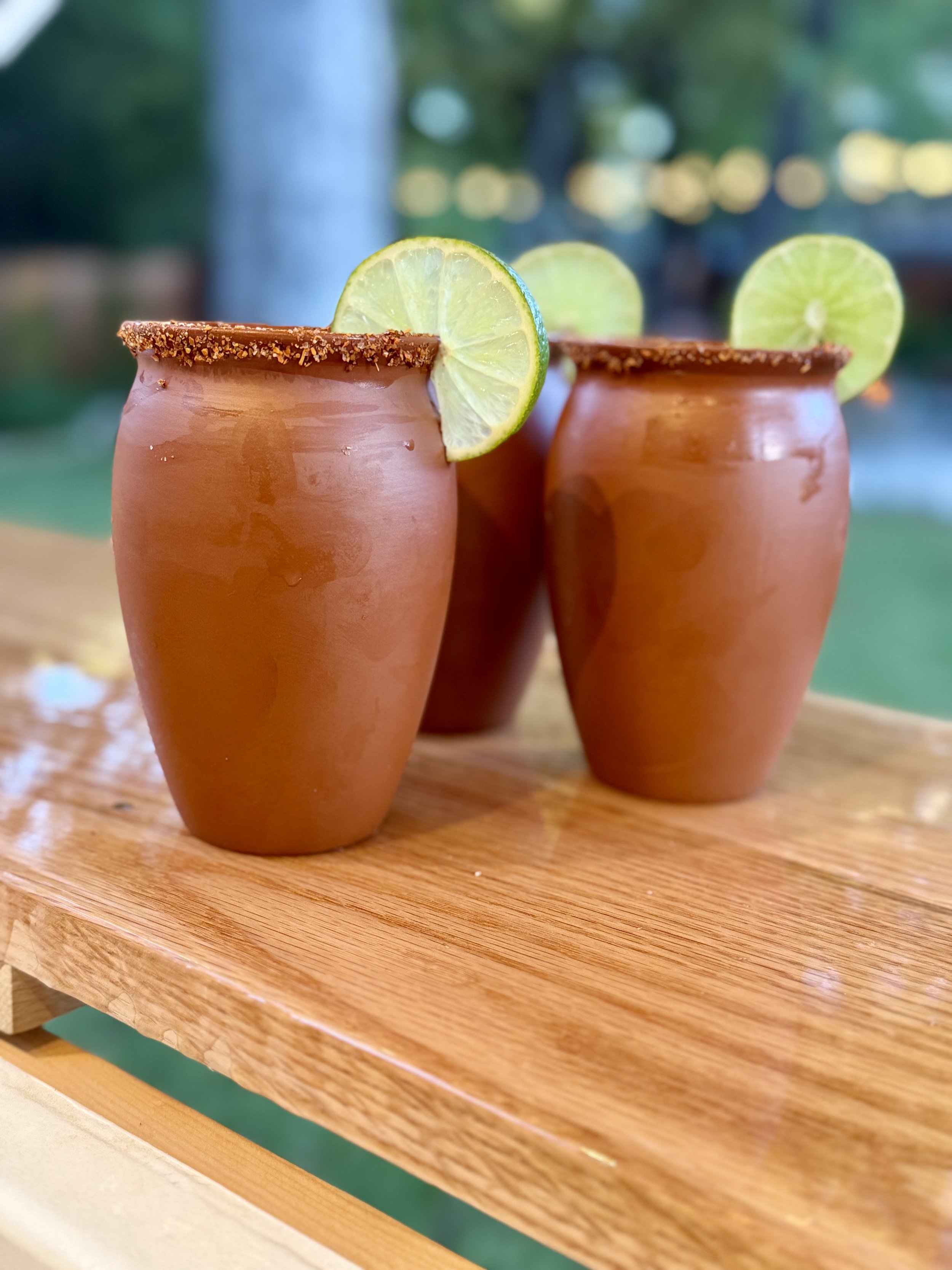 Three terracotta-colored glasses with salted rims and lime slices on the rim, sitting on a wooden surface, outdoors with blurred trees in the background.