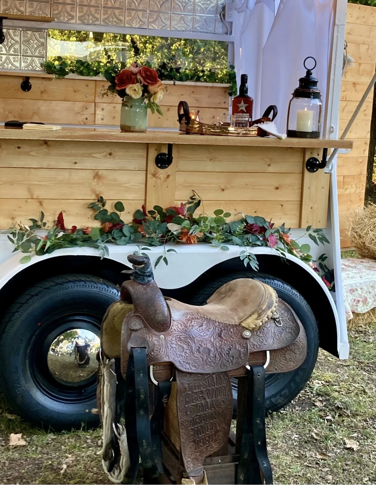 A rustic, vintage-style scene featuring a wooden food or drink kiosk built on a large tire wheel. Decorated with a floral arrangement on top of the wheel with green leaves and red, pink, and white flowers. In front of the setup is an elaborately tool