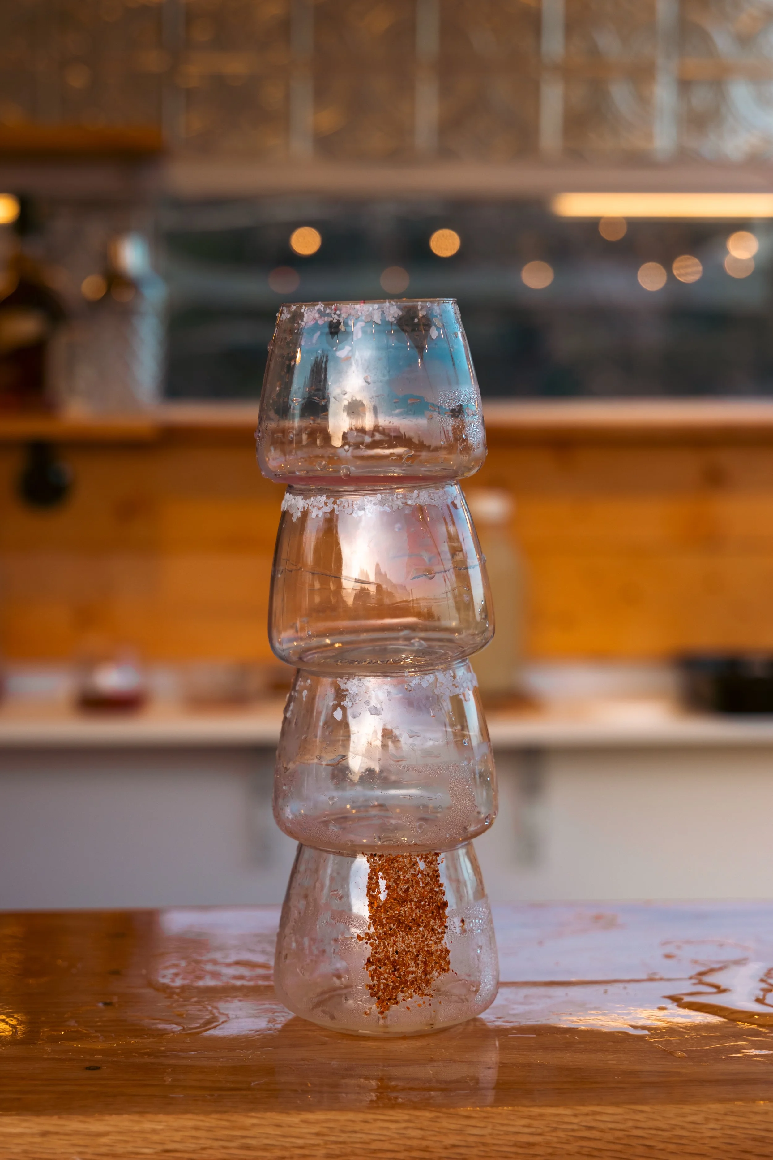 Stack of upside-down glasses with salt on the rims, forming a tower on a wooden table with spilled liquid around.