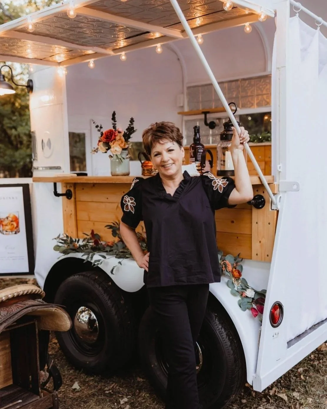 A woman standing in front of a decorated white mobile food cart, smiling and holding a pole. The cart has a wooden counter, flower arrangements, and is lit with string lights at dusk outdoors.