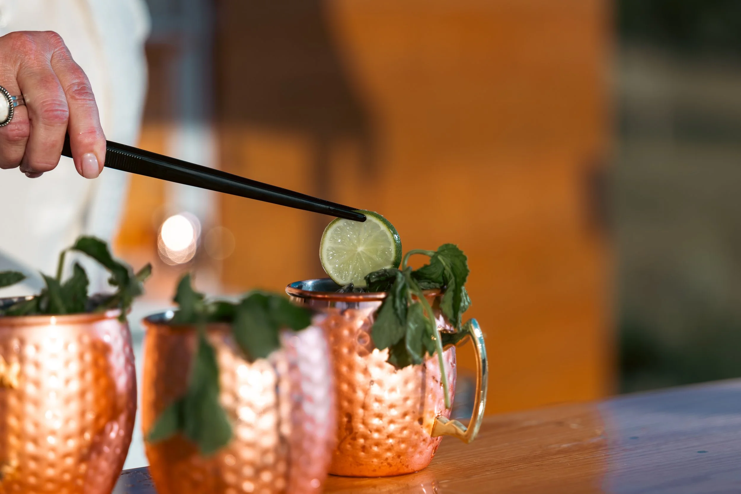 Close-up of a hand holding black tongs, gripping a lime slice over copper mugs filled with a drink garnished with mint leaves.