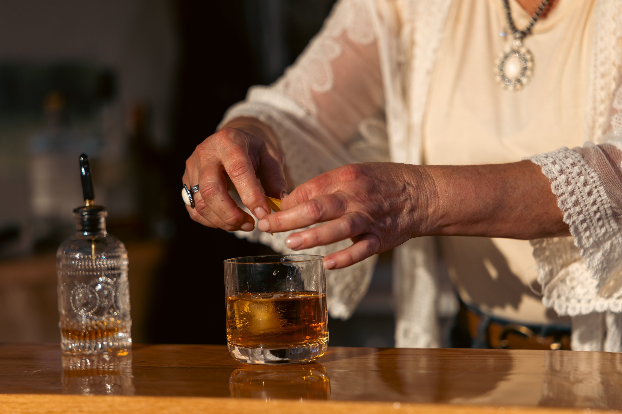 A person wearing a cream-colored, lace-embellished blouse adds a lemon twist to a glass of amber-colored liquid, with a water bottle nearby.