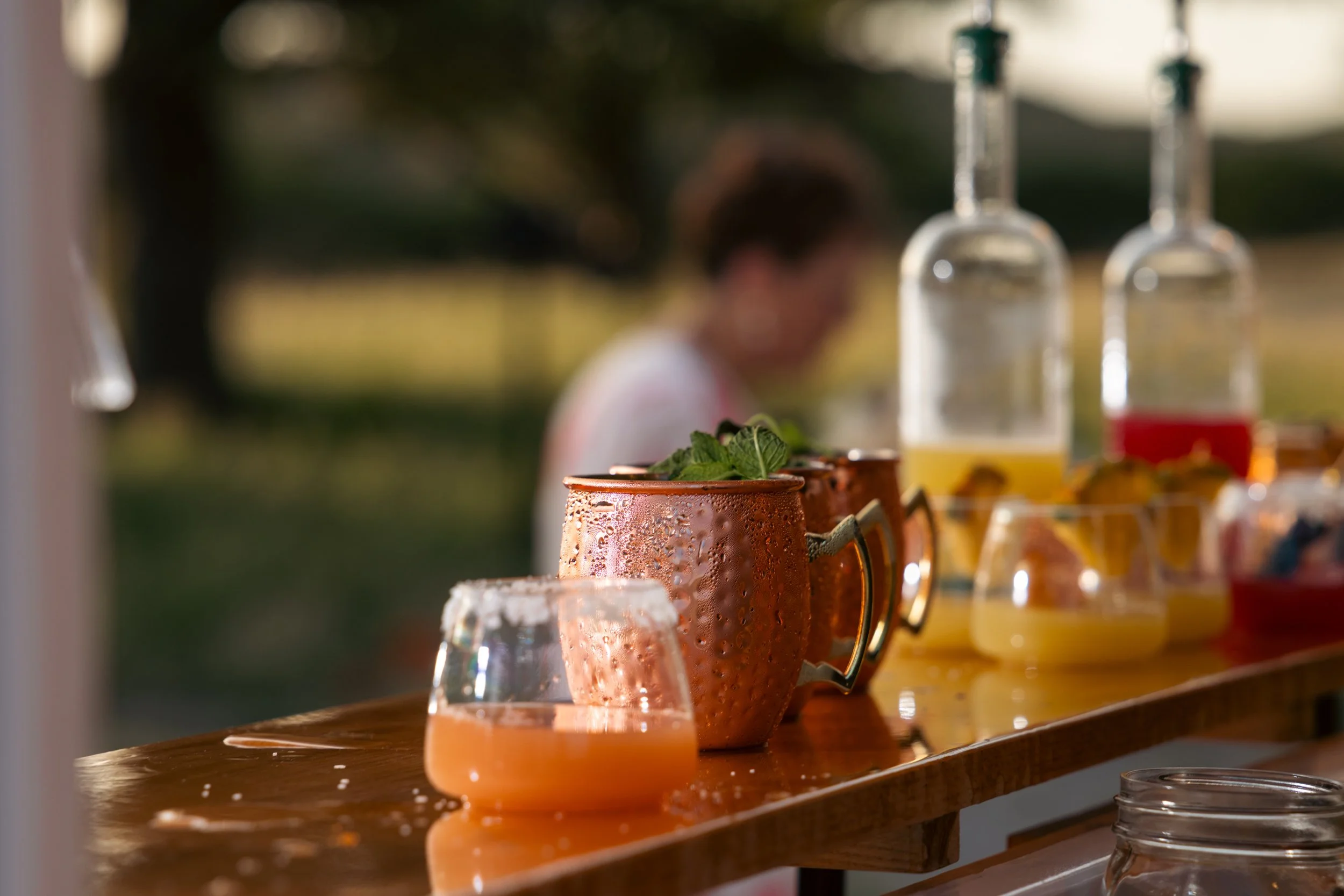 A row of containers and glasses with drinks on a wooden surface, with a blurred person and outdoor background in the distance.