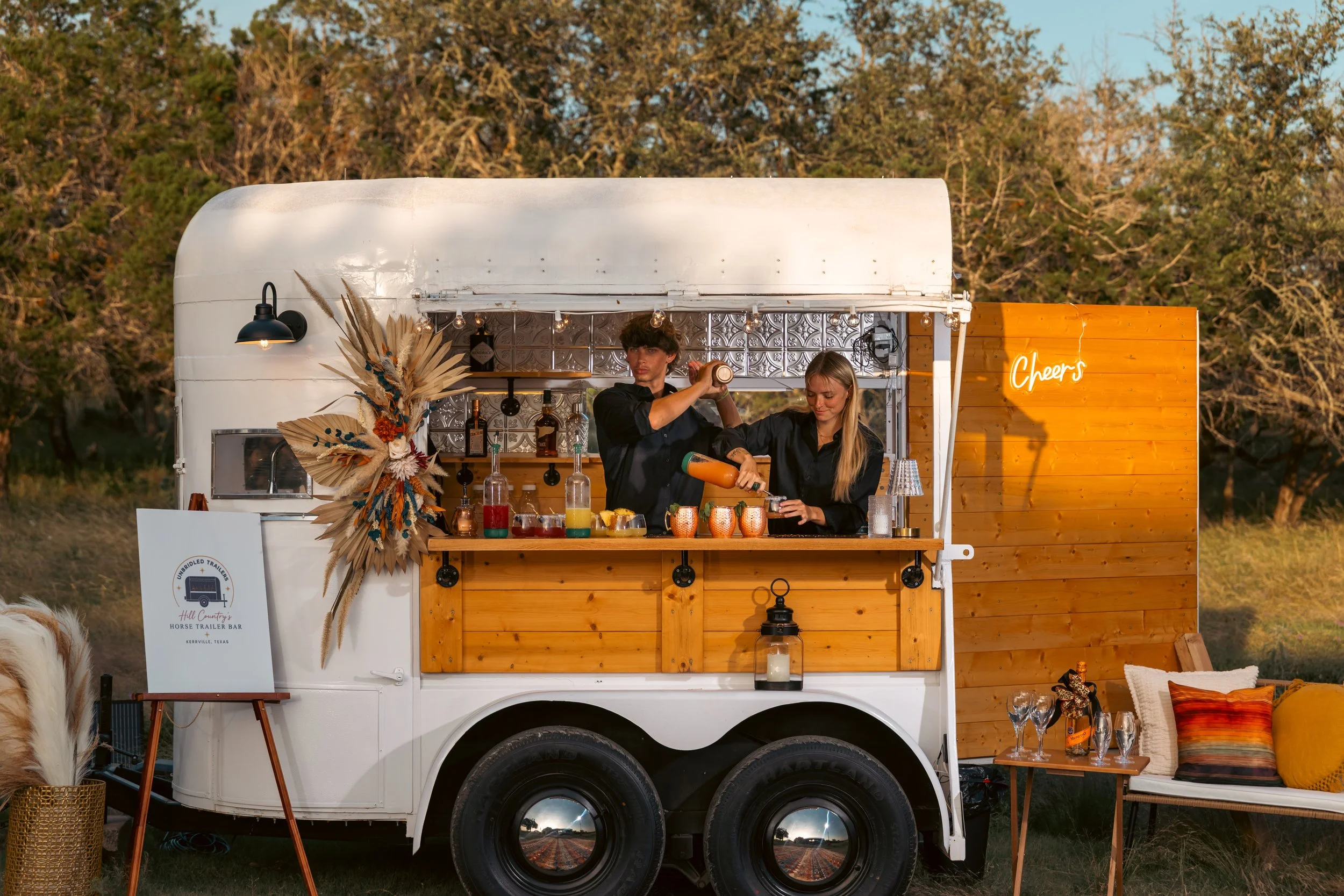 A mobile bar setup on a trailer with a wooden front, a small counter, and bar supplies, set outdoors during sunset with trees in the background. A person is preparing drinks while another assists, decorated with dried plants and a sign that says 'Cheers'.