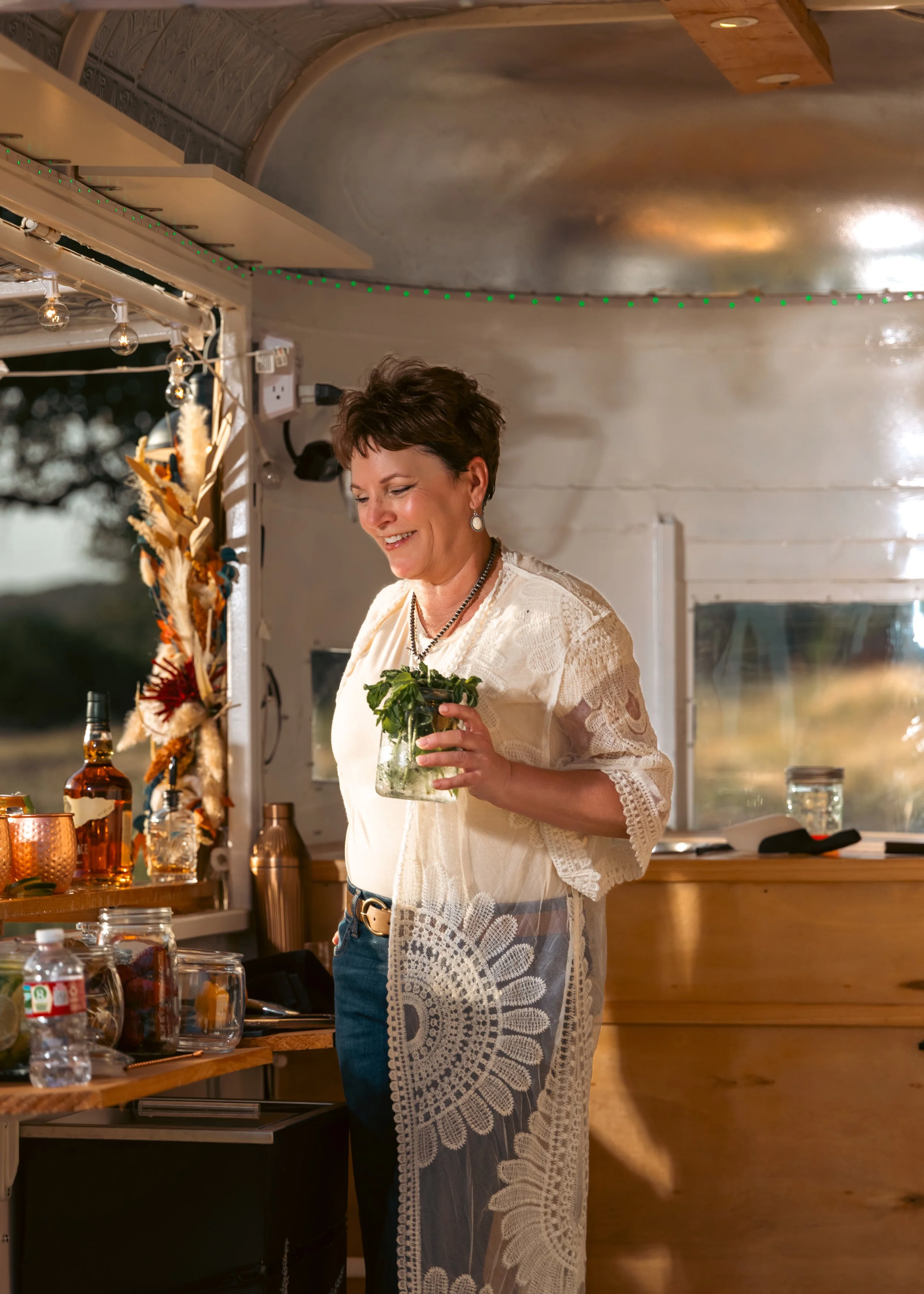 A woman standing inside a food truck, smiling, holding a small potted plant, with a bar area and various bottles and glasses nearby.