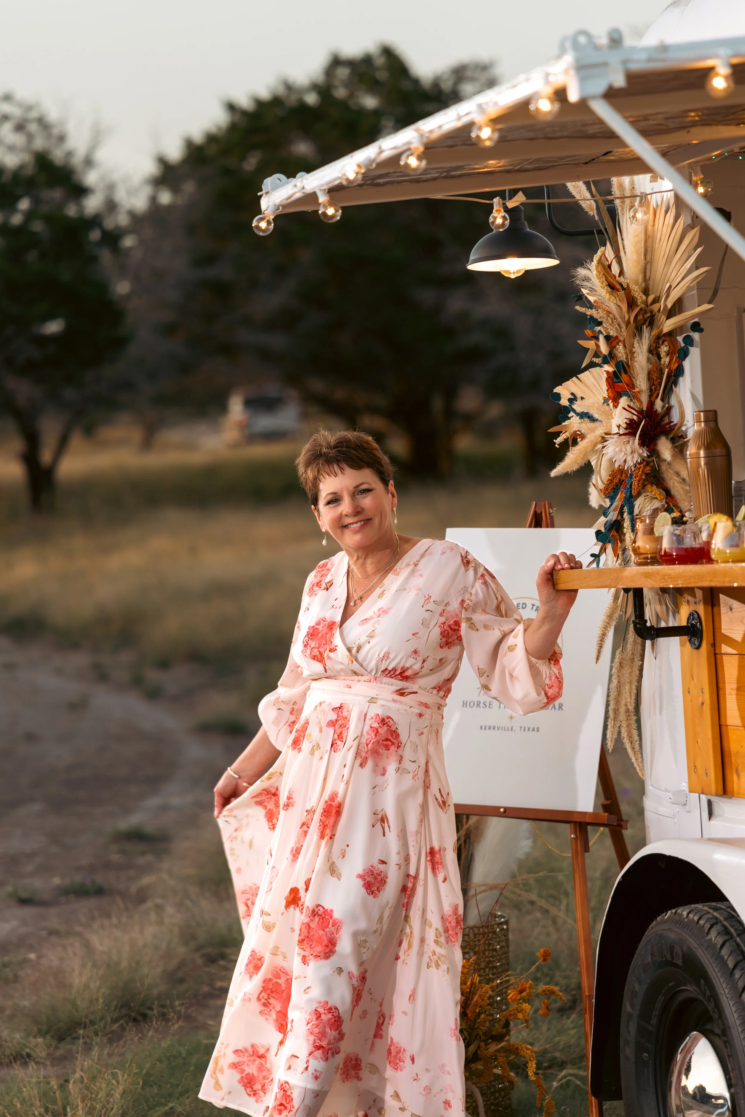 A woman in a floral pink and white dress standing outdoors at sunset, beside a food truck decorated with dried flowers and string lights.