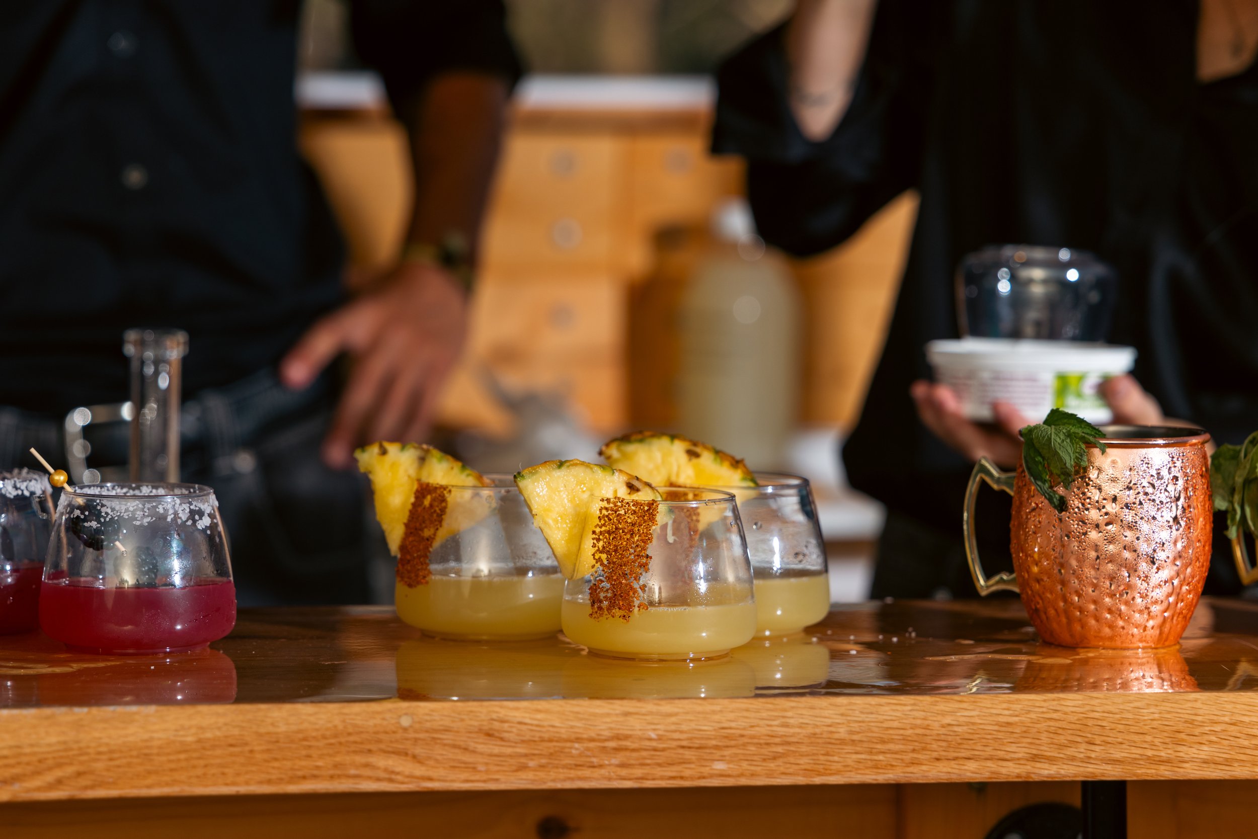 Several cocktails with pineapple slices and garnishes on a wooden bar counter, with people in the background at a social gathering.