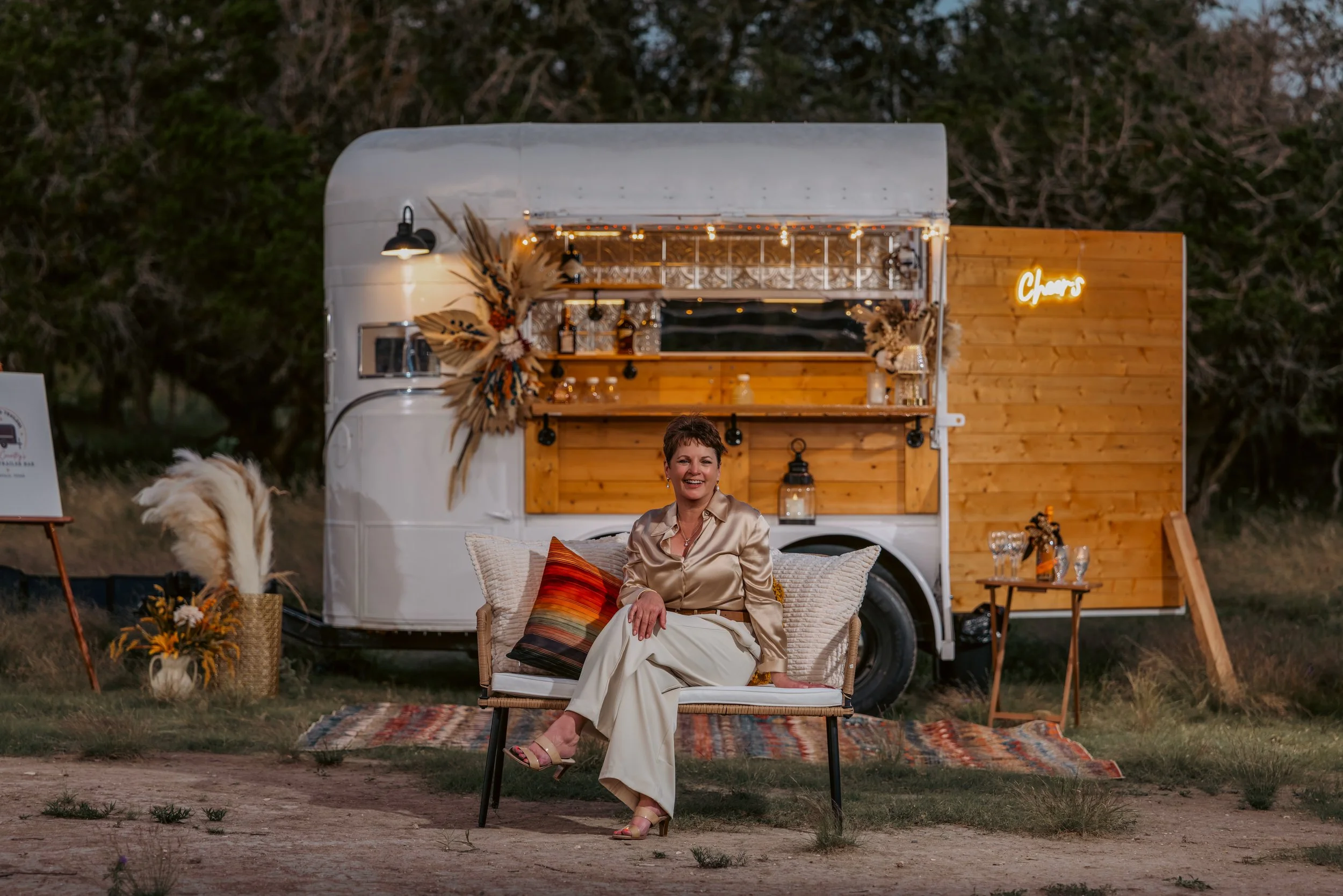 A woman sitting on a wicker chair in front of a vintage food truck decorated with dried flowers and string lights. She is smiling and wearing a beige satin blouse, cream-colored pants, and pink heels. There is a colorful patterned rug on the ground and a small table with glasses nearby. The scene is outdoors at dusk with trees in the background.