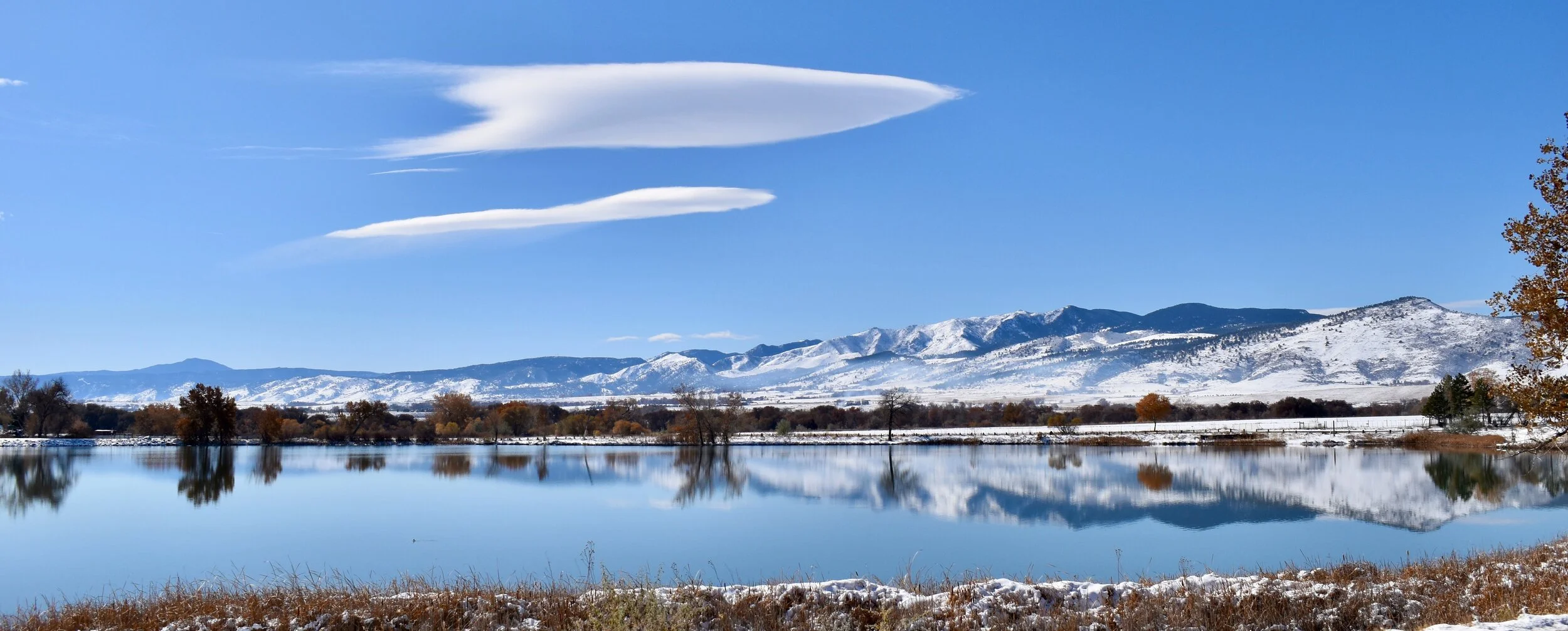 Snow-covered mountains with a clear blue sky, a few unique lenticular clouds, a reflective body of water in the foreground, and some trees along the shoreline.
