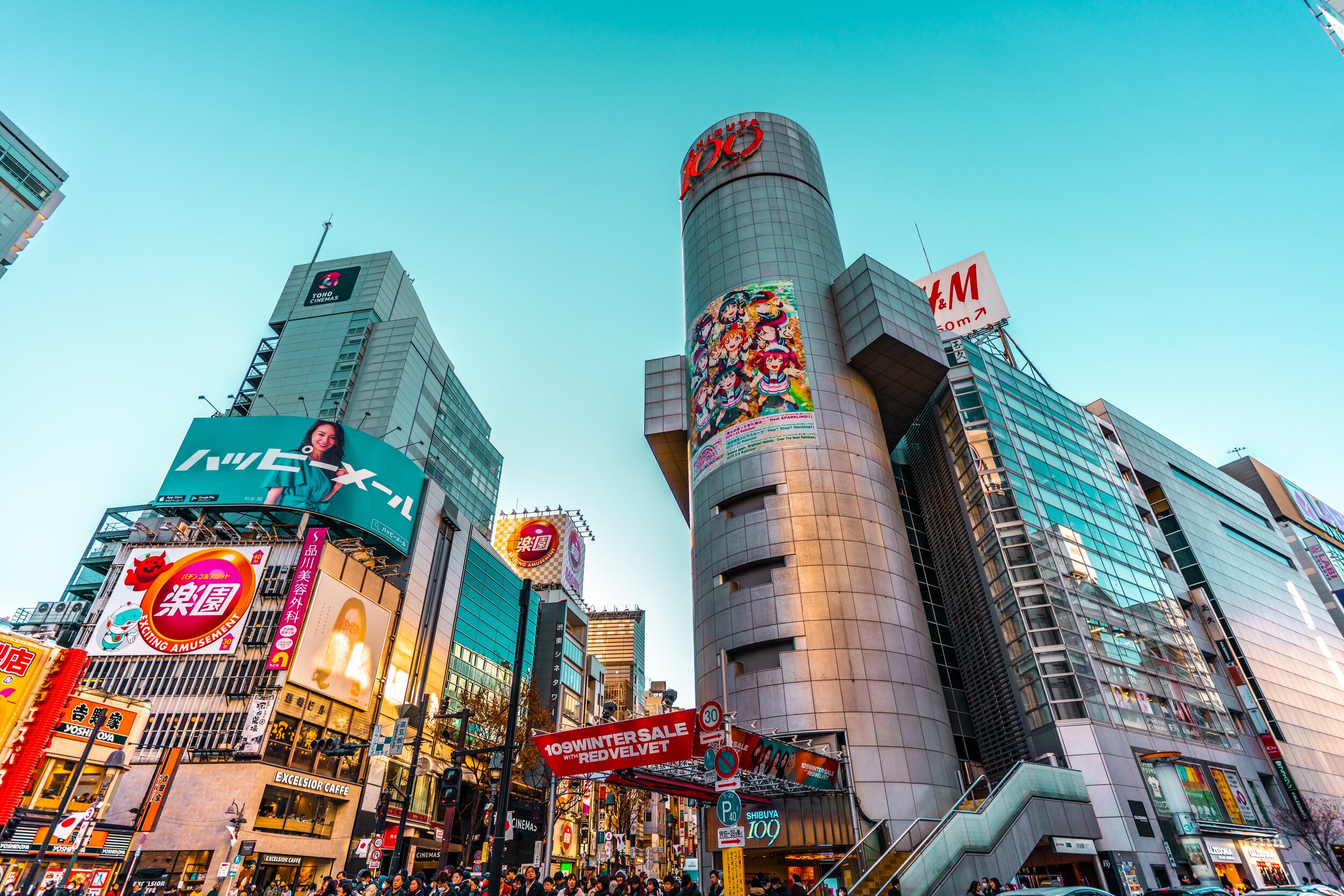Cityscape of Shibuya with tall buildings, billboards, advertisements, and a crowd of people on the street, during daytime.