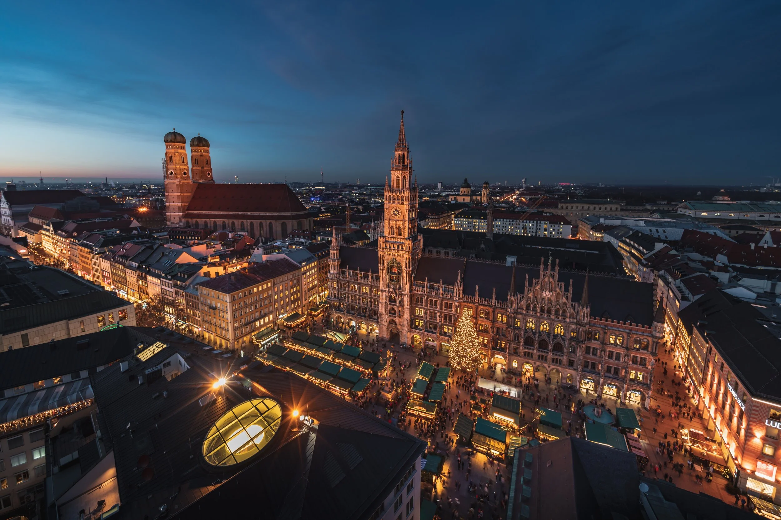 Aerial view of Munich's Christmas market in the evening, featuring historic buildings, a large decorated Christmas tree, and market stalls with lights.
