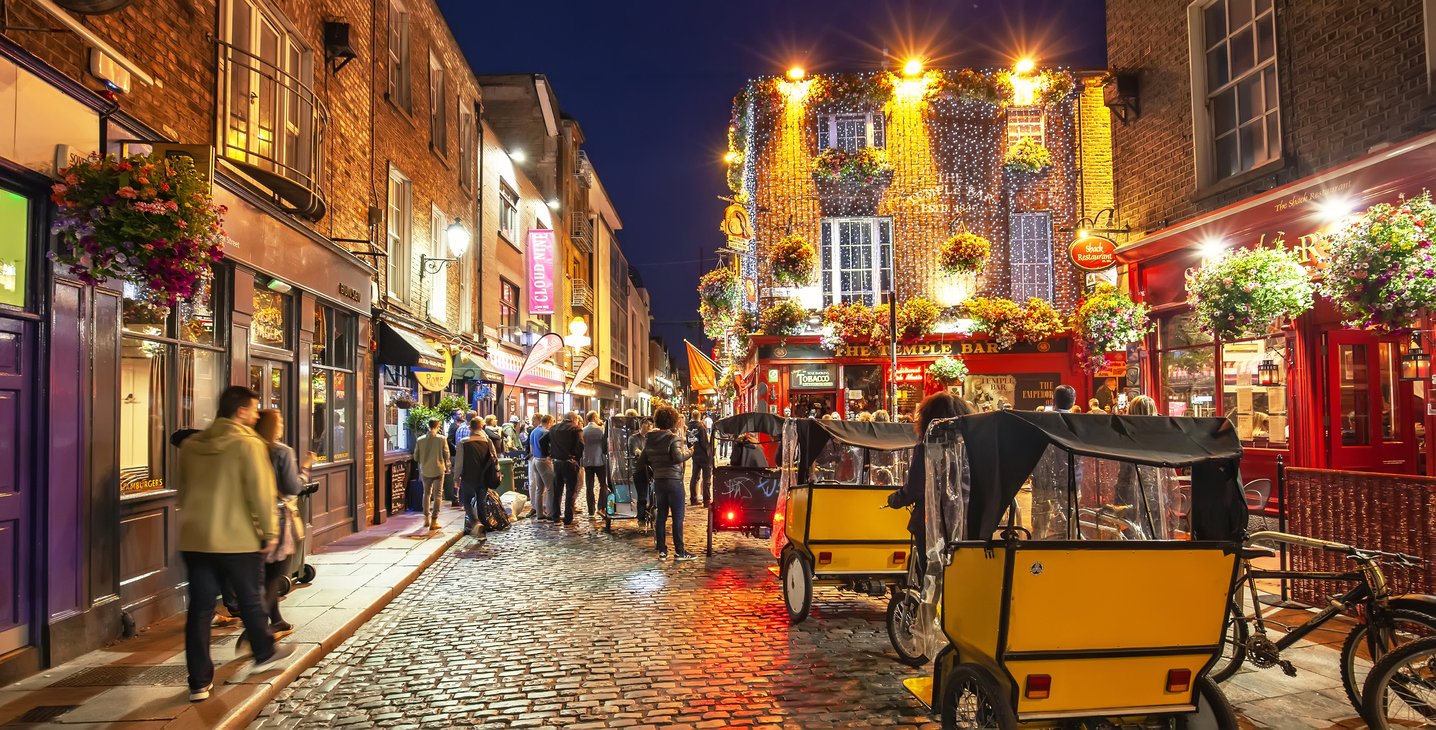A lively street scene at night with colorful shops, hanging flower baskets, and outdoor seating. Pedestrians walk along the cobblestone street, some carrying umbrellas. Bright streetlights and decorative lights illuminate the area, creating a vibrant