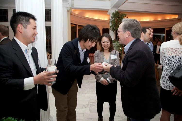 A group of people at a social event, with one person handing something to another while others look on, standing indoors with warm lighting and plants in the background.