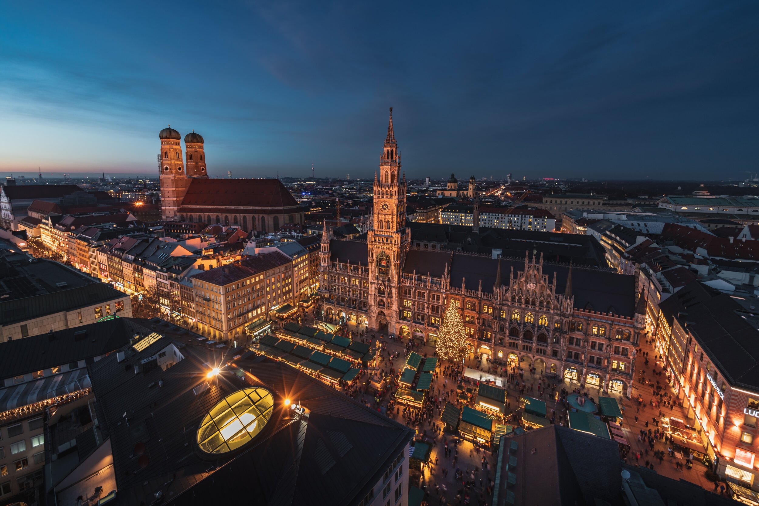 Aerial view of Munich's Marienplatz at dusk, featuring the New Town Hall with its clock tower, a Christmas tree in the square, and markets with stalls, surrounded by historic buildings and a darkening sky.