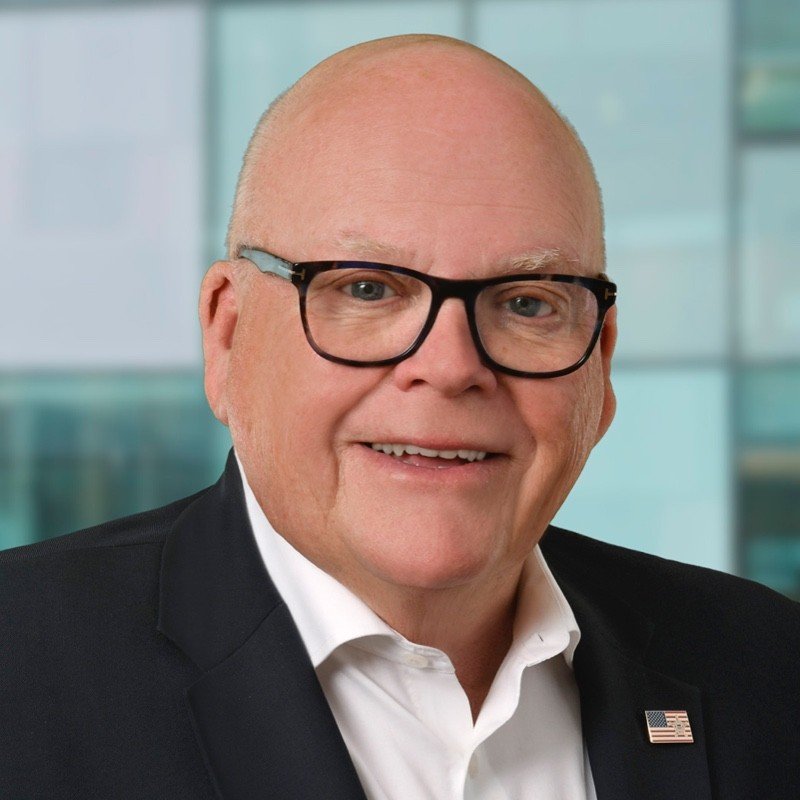 A middle-aged man with glasses and a bald head, wearing a black suit, white shirt, and an American flag pin, smiling in front of a modern glass building.
