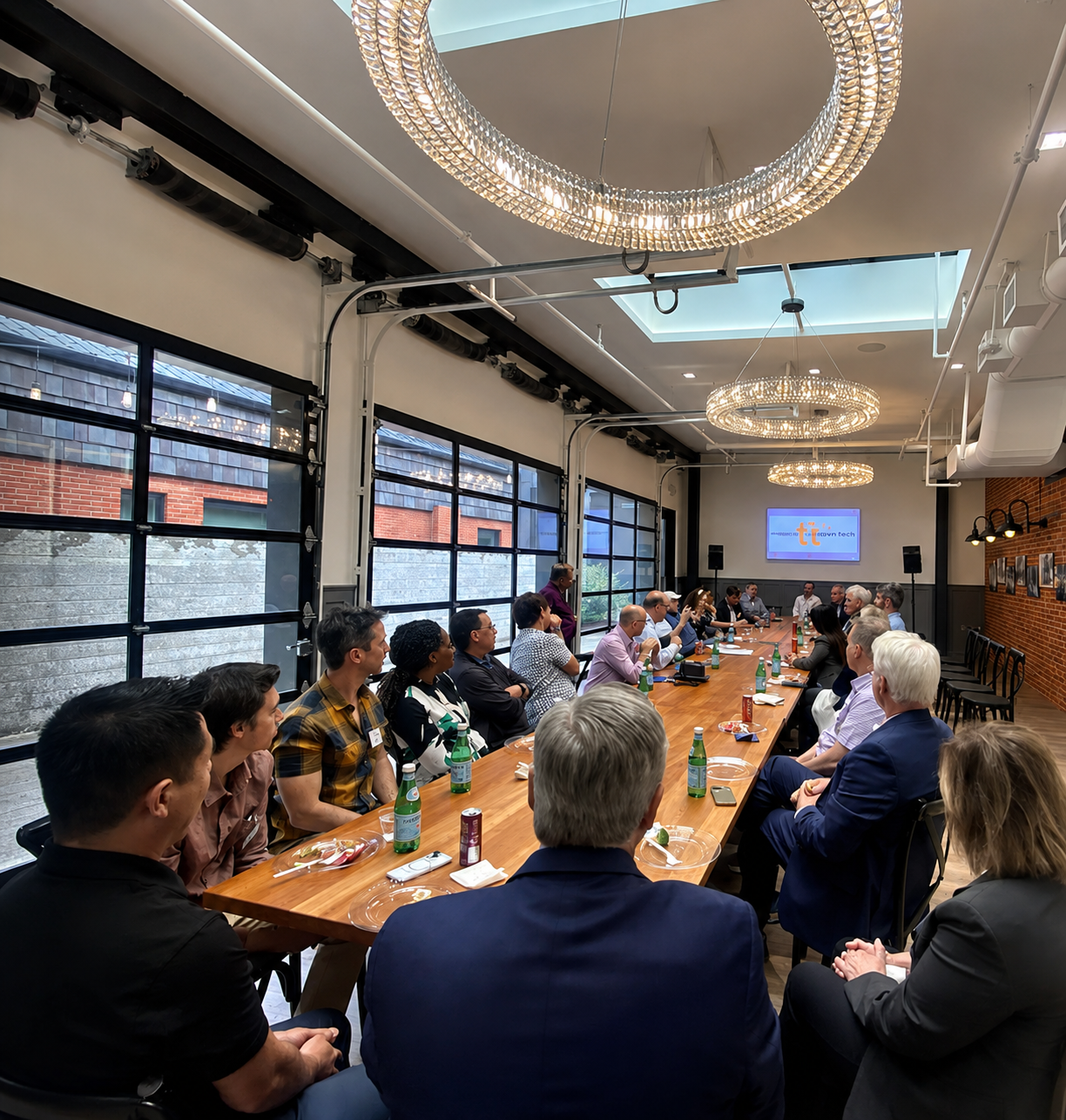 People seated around a long conference table in a modern meeting room with large windows, a presentation screen, and decorative ceiling chandeliers.