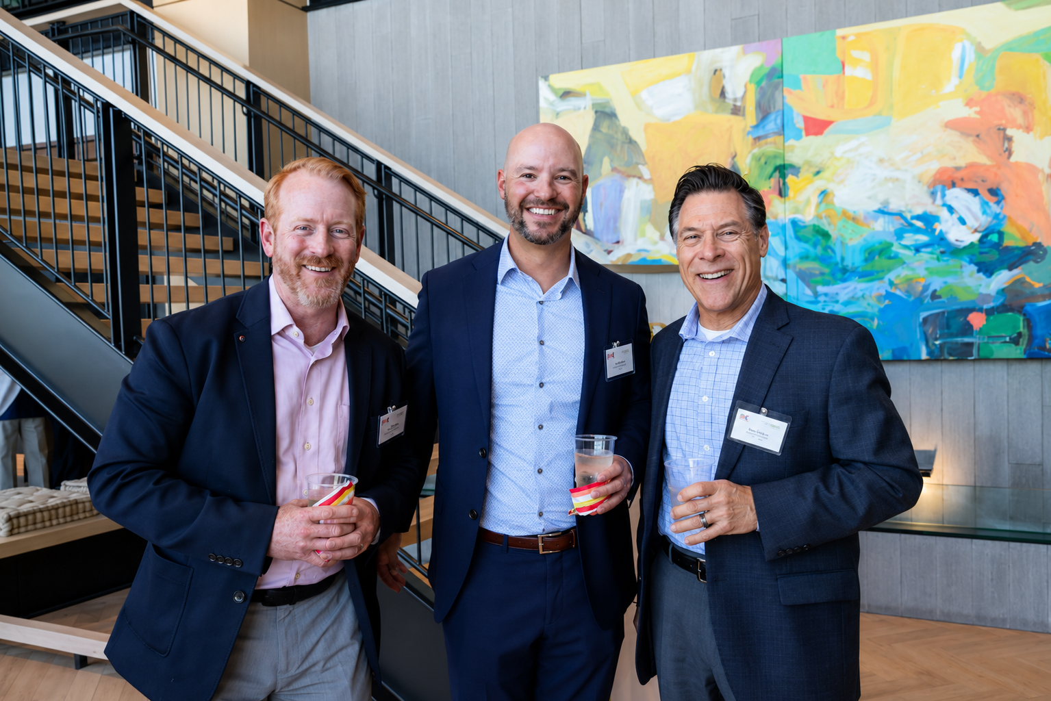 Three men in business suits smiling and holding drinks at a corporate event, standing in front of colorful abstract artwork.