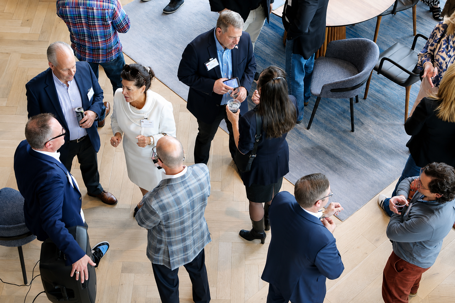 A group of people at a professional networking event, standing and talking in a room with wooden floors and a carpeted area, some holding drinks and wearing business attire.