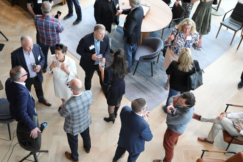A group of professionals at a networking event engaging in conversations. Some are standing in circles, others are sitting or walking around, with most holding drinks or smartphones. The setting is indoors with wooden flooring and modern furniture.
