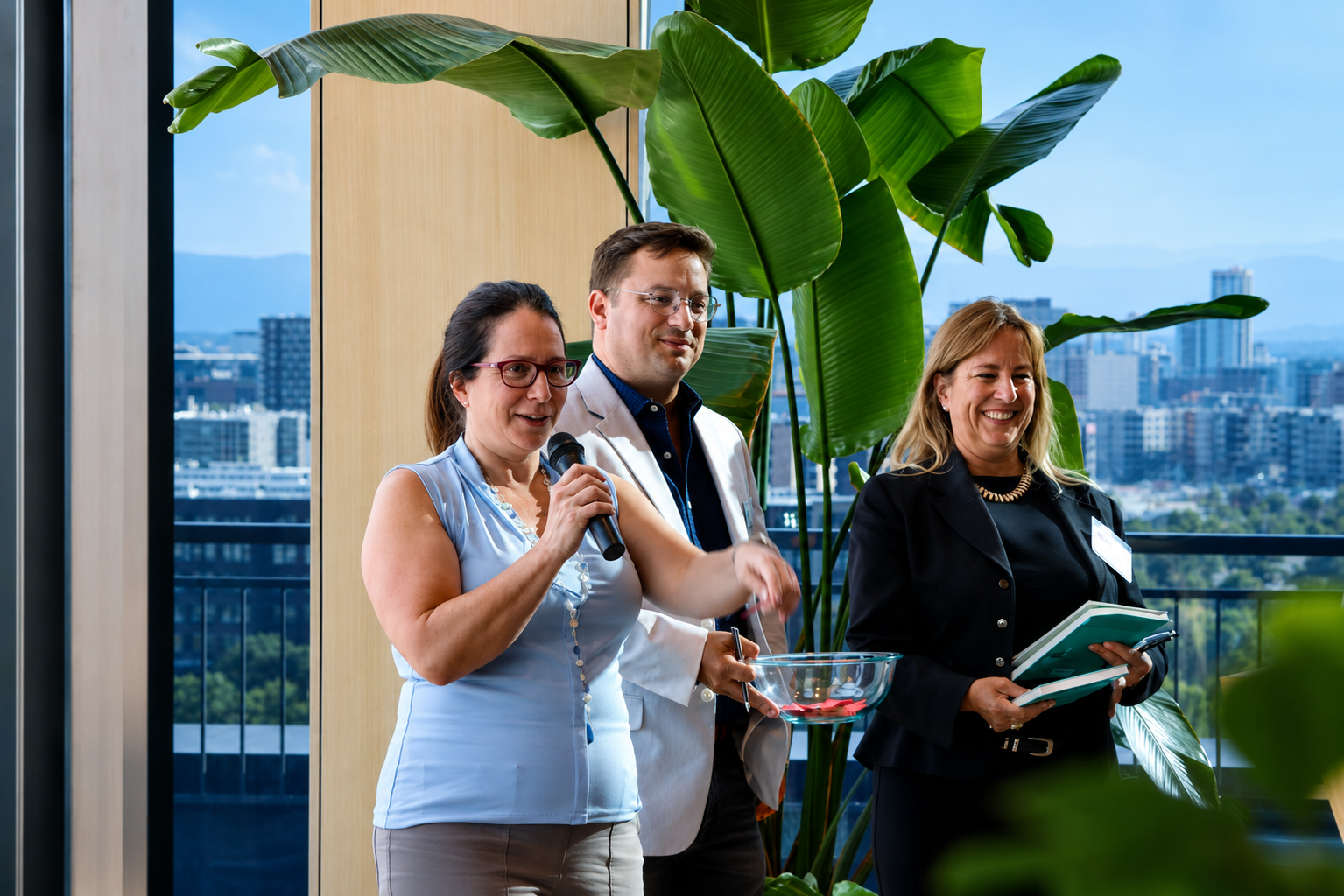 Three professionals standing near large green indoor plants, with city buildings visible through a window behind them. The woman on the left, wearing glasses and a light blue sleeveless top, is speaking into a microphone. The man in the middle, dressed in a white blazer and blue shirt, is holding a bowl. The woman on the right, wearing a black blazer and gold necklace, is holding a notebook and pen, smiling.