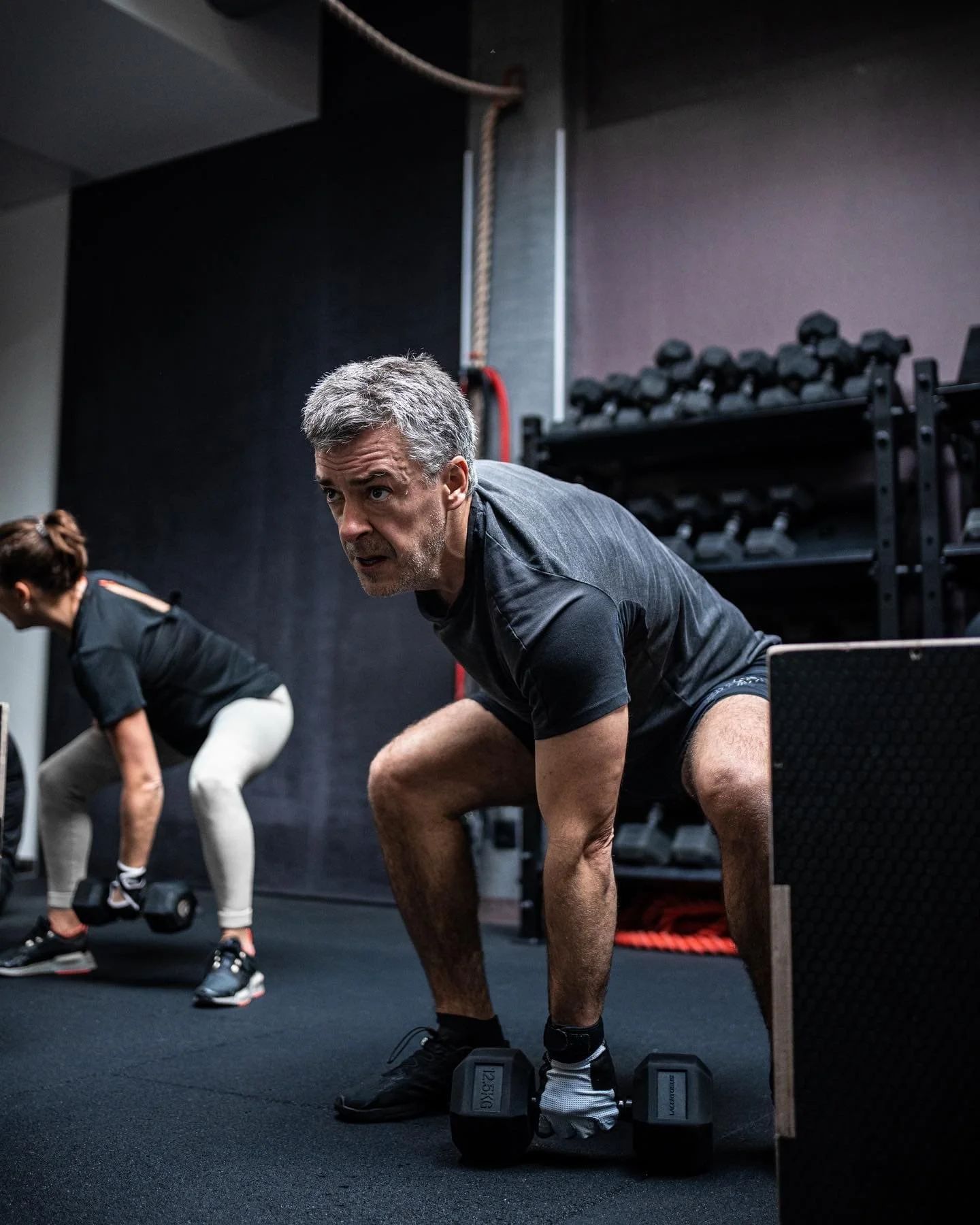 Deux personnes, un homme et une femme, font de l'exercice de squat avec des haltères dans une salle de sport en intérieur, avec des racks de poids en arrière-plan.