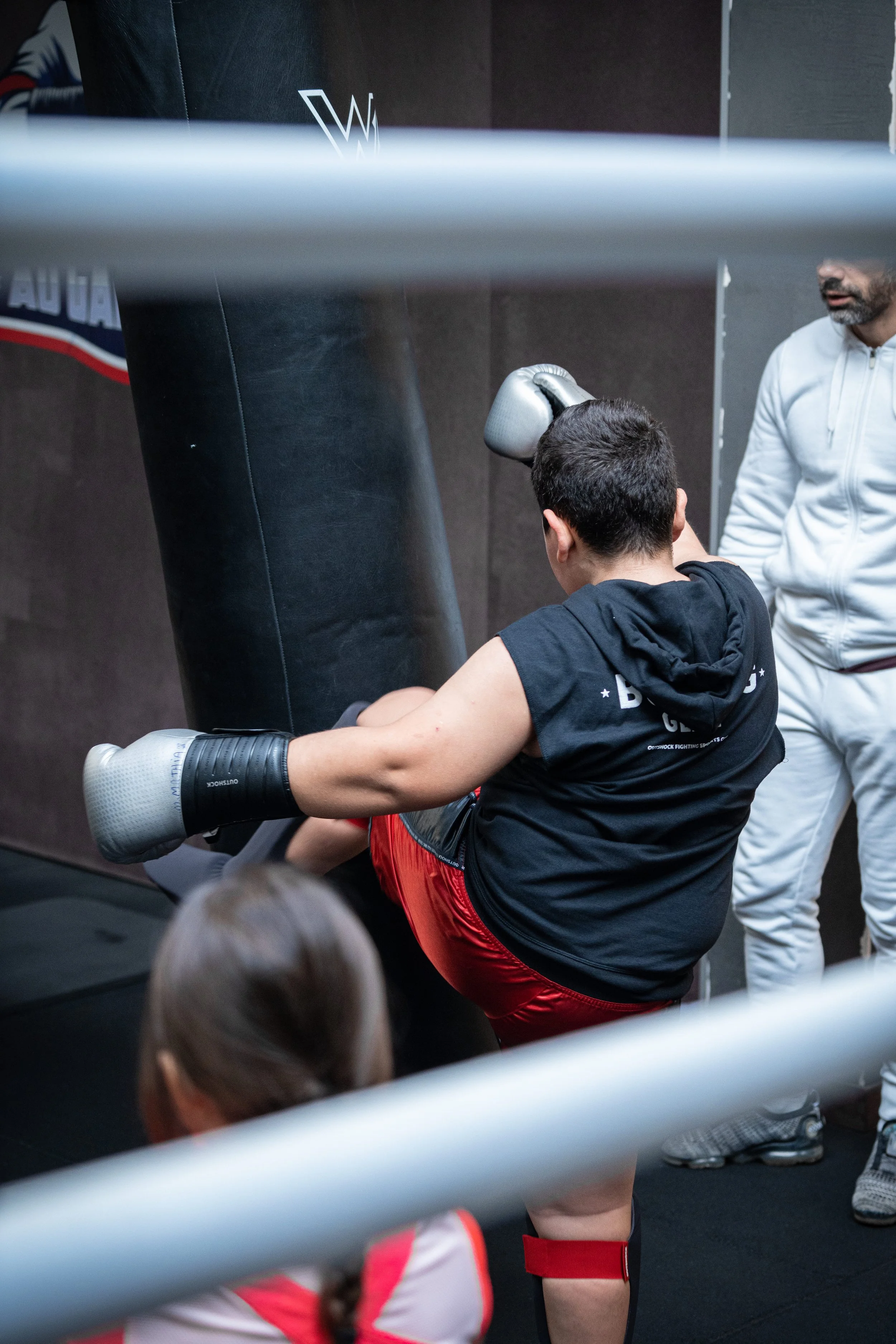 Un homme en t-shirt noir avec un short rouge, portant des gants de boxe, effectue un coup de pied latéral sur un sac de frappe dans un gymnase, avec deux personnes en arrière-plan, dont une en survêtement blanc.