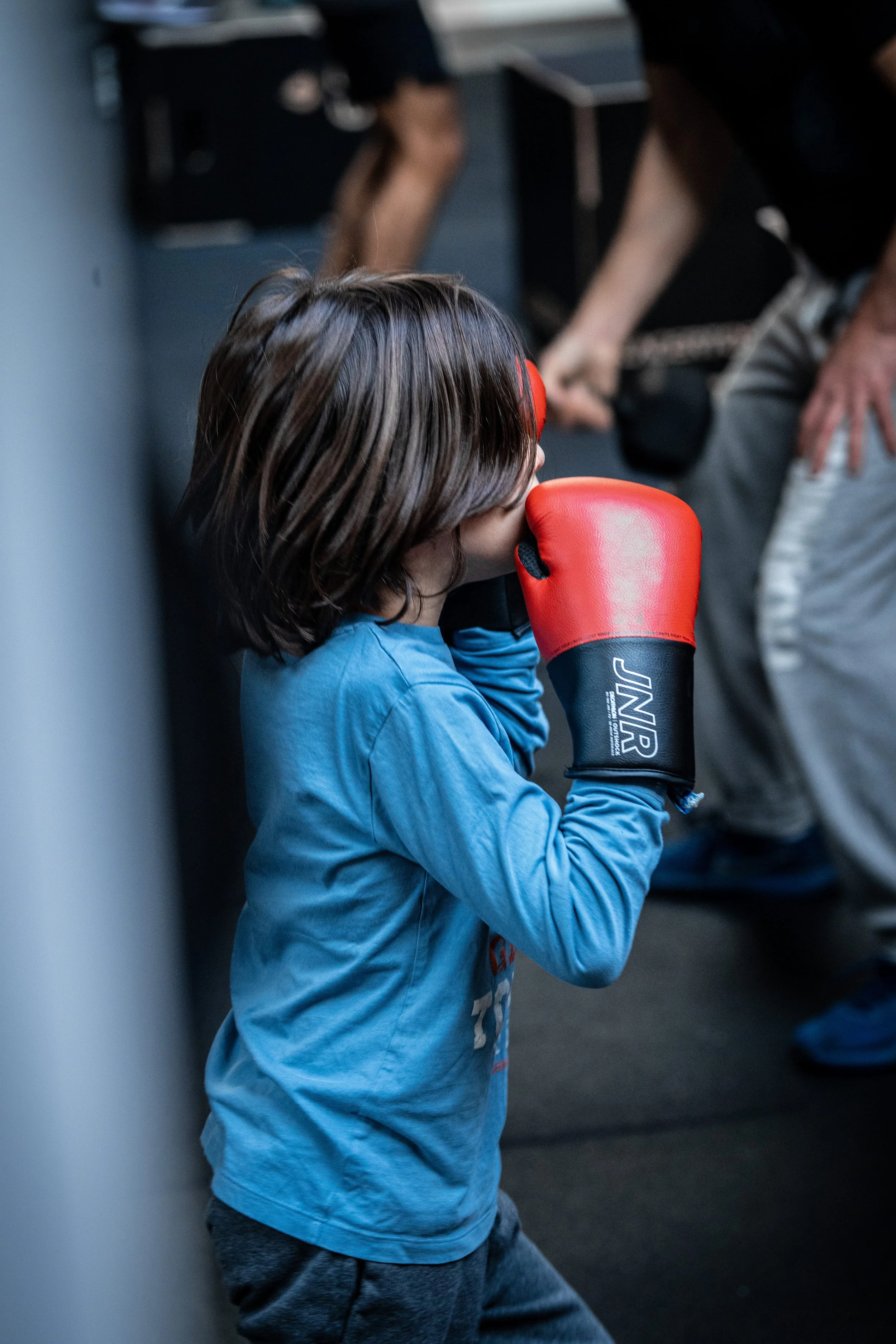 Un jeune garçon avec des gants de boxe rouges, en position de garde, dans une salle d'entraînement ou de compétition, avec des adultes en arrière-plan.