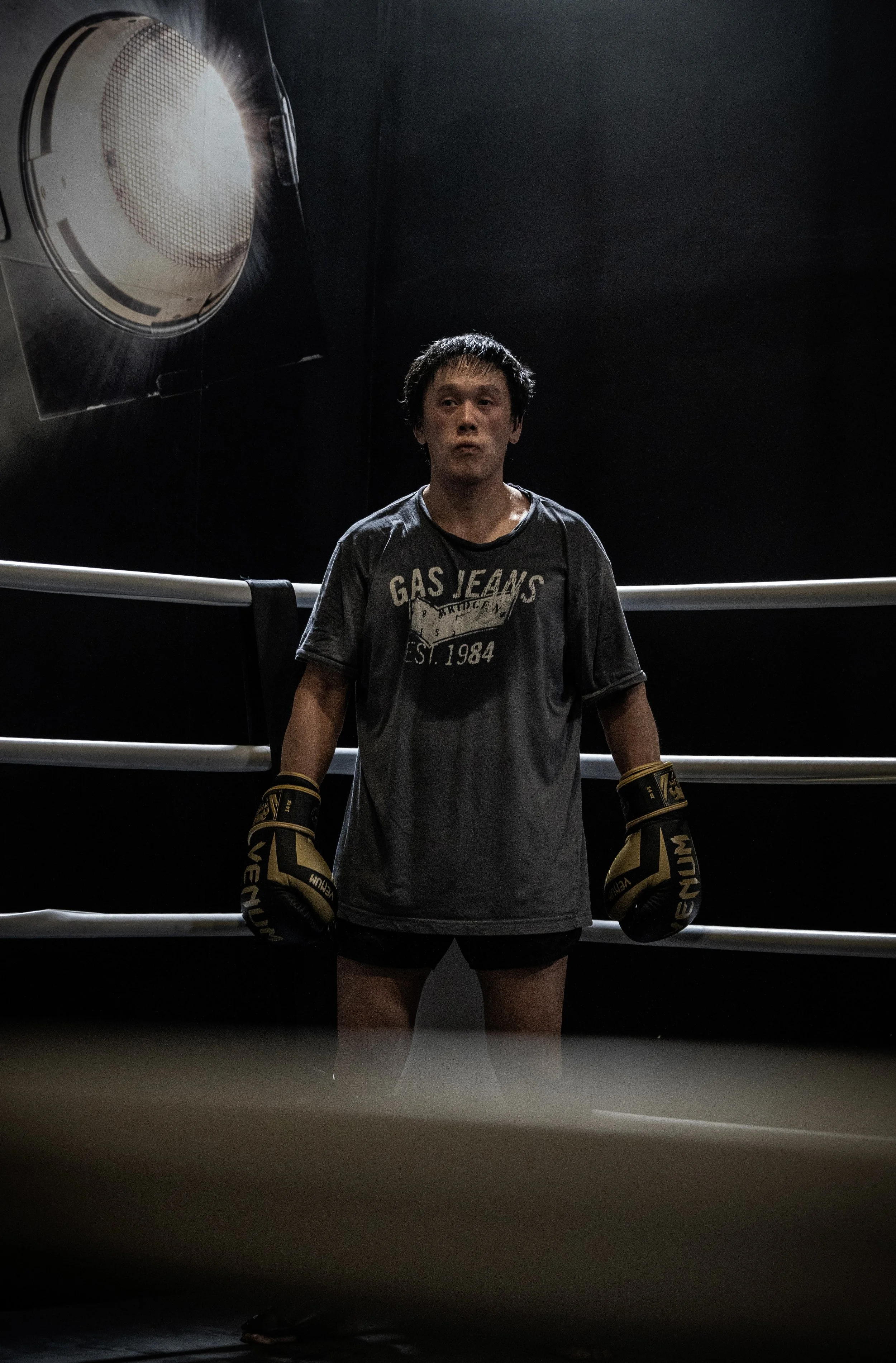 Un homme en tenue de boxe portant des gants de boxe noirs et dorés, debout dans un ring de boxe sombre.