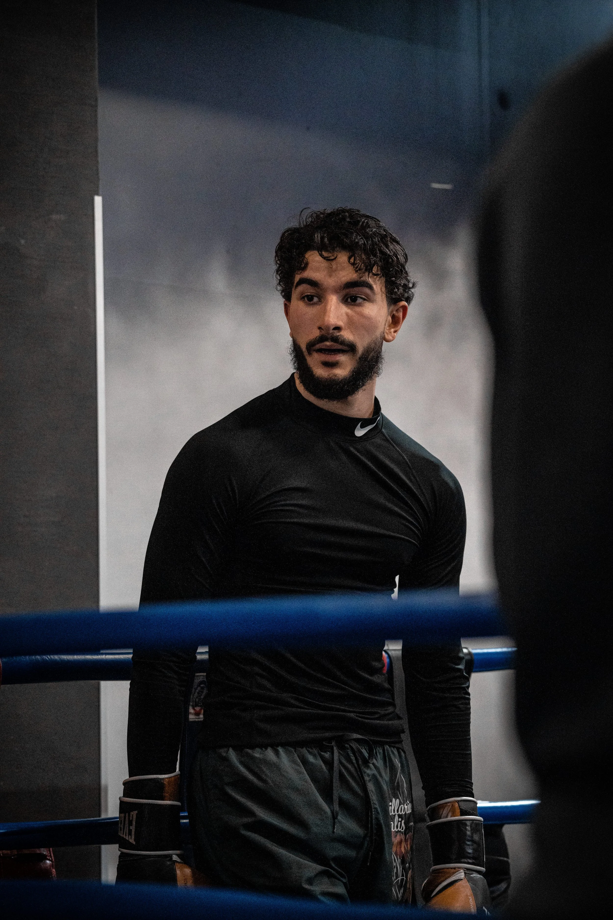 Un homme avec une barbe et des cheveux bouclés, portant une tenue de boxe noire, est debout dans un ring de boxe, regardant vers la gauche.