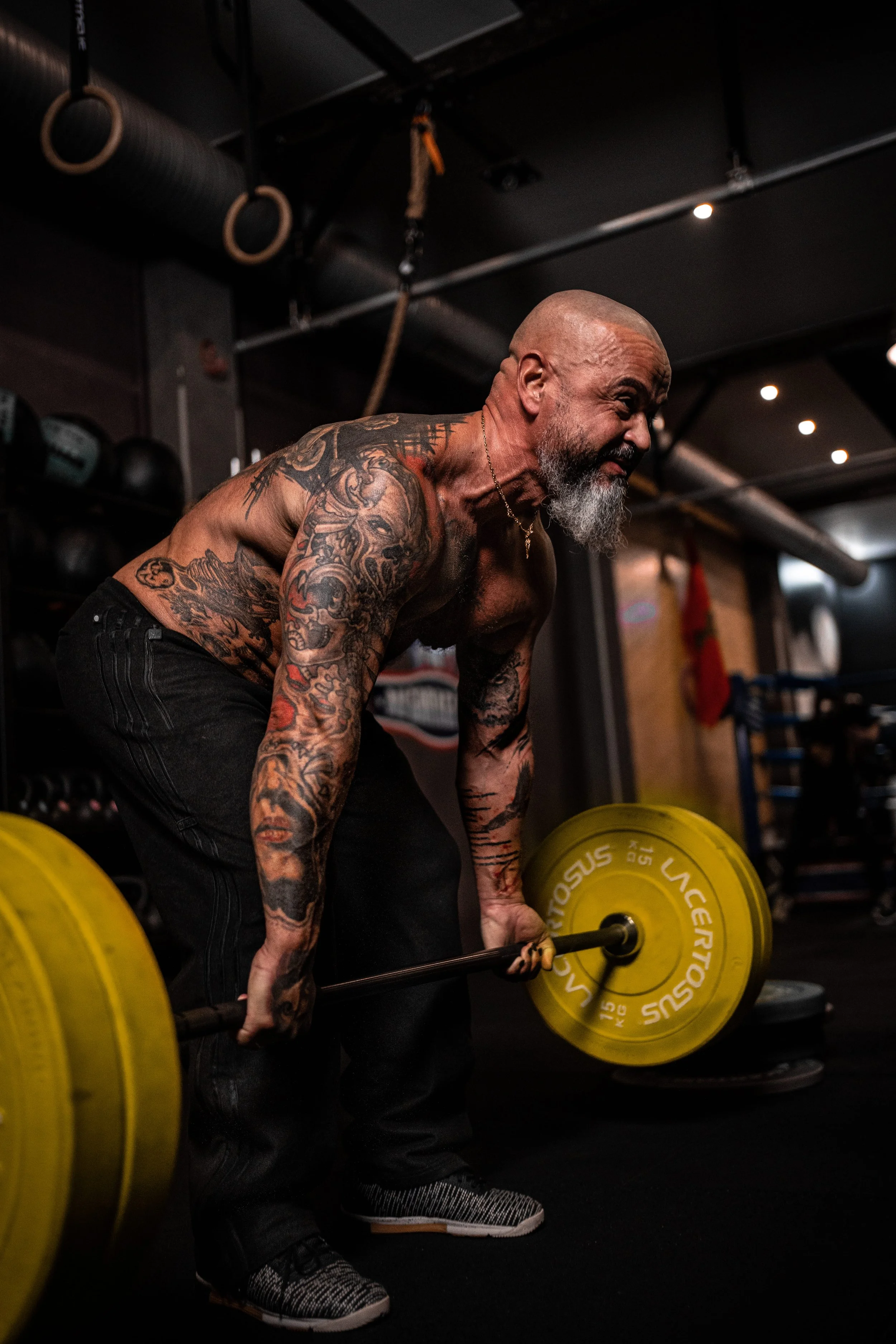 Un homme musclé et tatoué, avec une barbe grise, en train de faire un deadlift avec une barre chargée de poids jaunes dans une salle de sport sombre.