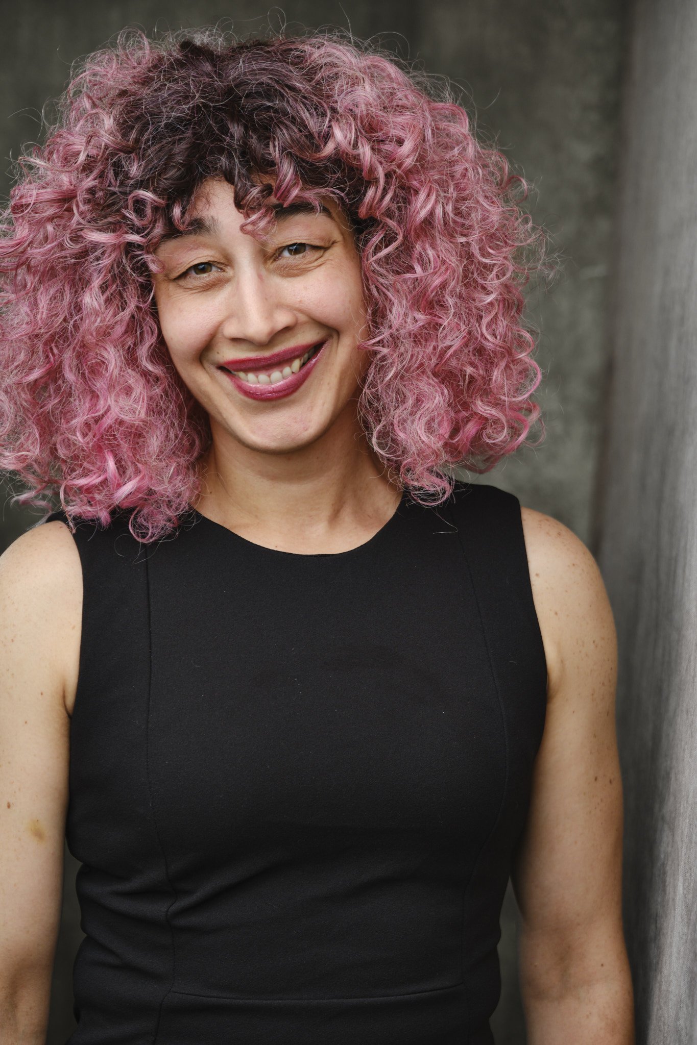 A woman with curly hair that is dark at the roots and pink at the tips, smiling while wearing a sleeveless black top, standing outdoors near a tree.