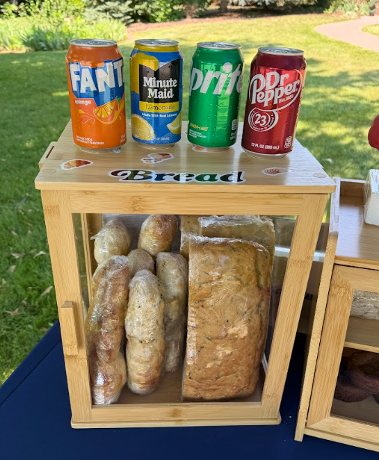 Four cans of soda and lemonade on top of a small wooden bread storage box with a glass door showing loaves of bread inside, outdoors on a sunny day.