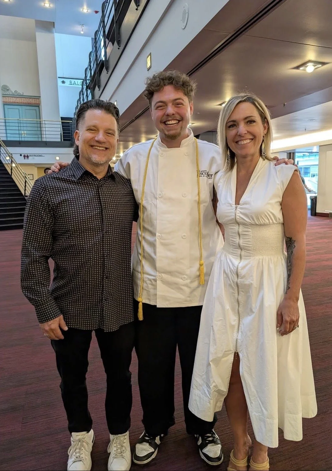 Three people standing together indoors, smiling for a photo. The person in the middle is wearing a chef's coat with a yellow rope, and the other two are dressed in casual and dressy attire.