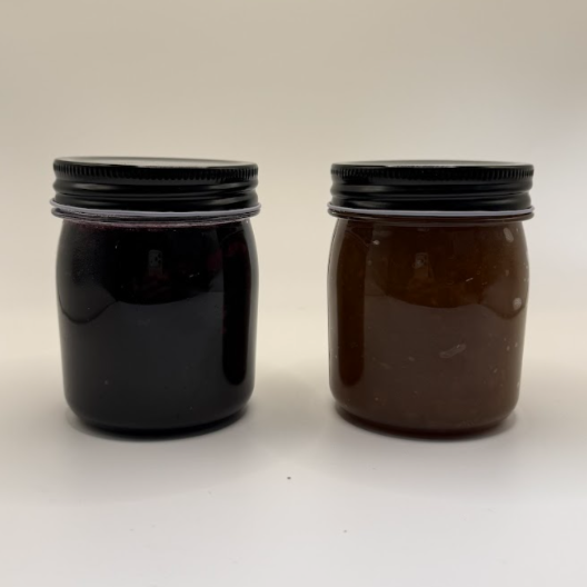 Two small glass jars with black screw-on lids, one filled with dark-colored jam or jelly and the other with brown-colored jam or jelly, placed on a white surface against a plain background.