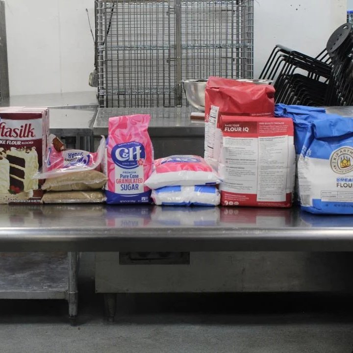 Assorted baking supplies on a stainless steel table, including flour bags, sugar, and baking powder, in a commercial kitchen or bakery setting.