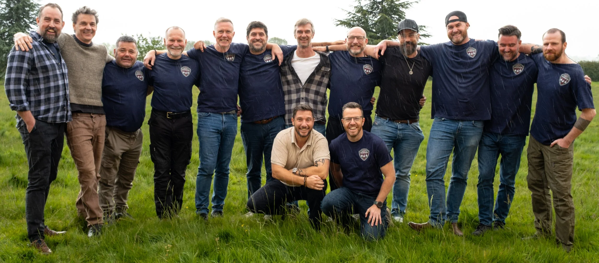 Group of men standing together outdoors on grass, smiling, with some wearing matching shirts with a logo.