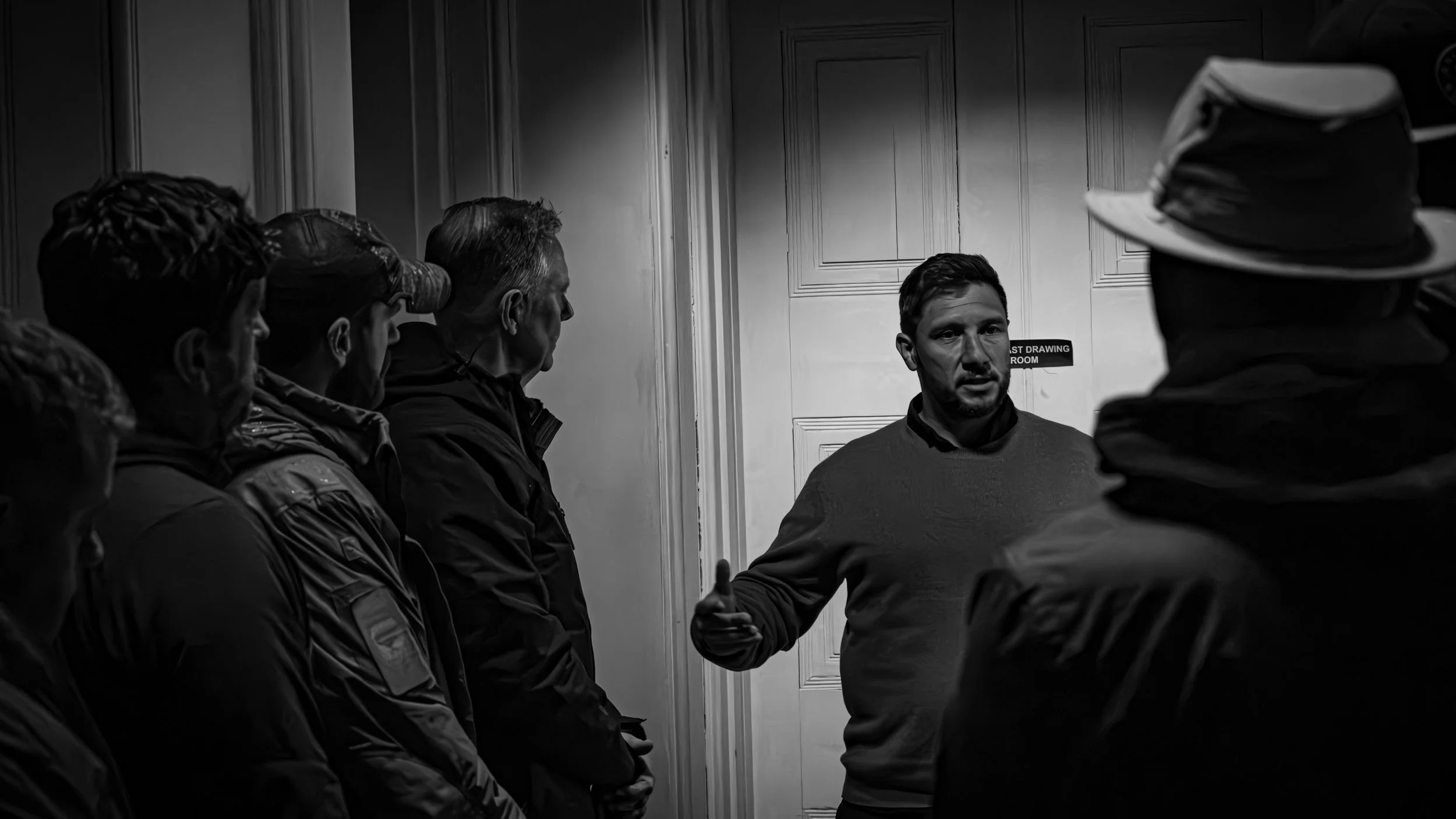 A man is speaking to a group of people in a small room with wooden paneling. The group appears to be listening attentively, with some taking notes or wearing hats. The setting suggests an informal meeting or discussion.