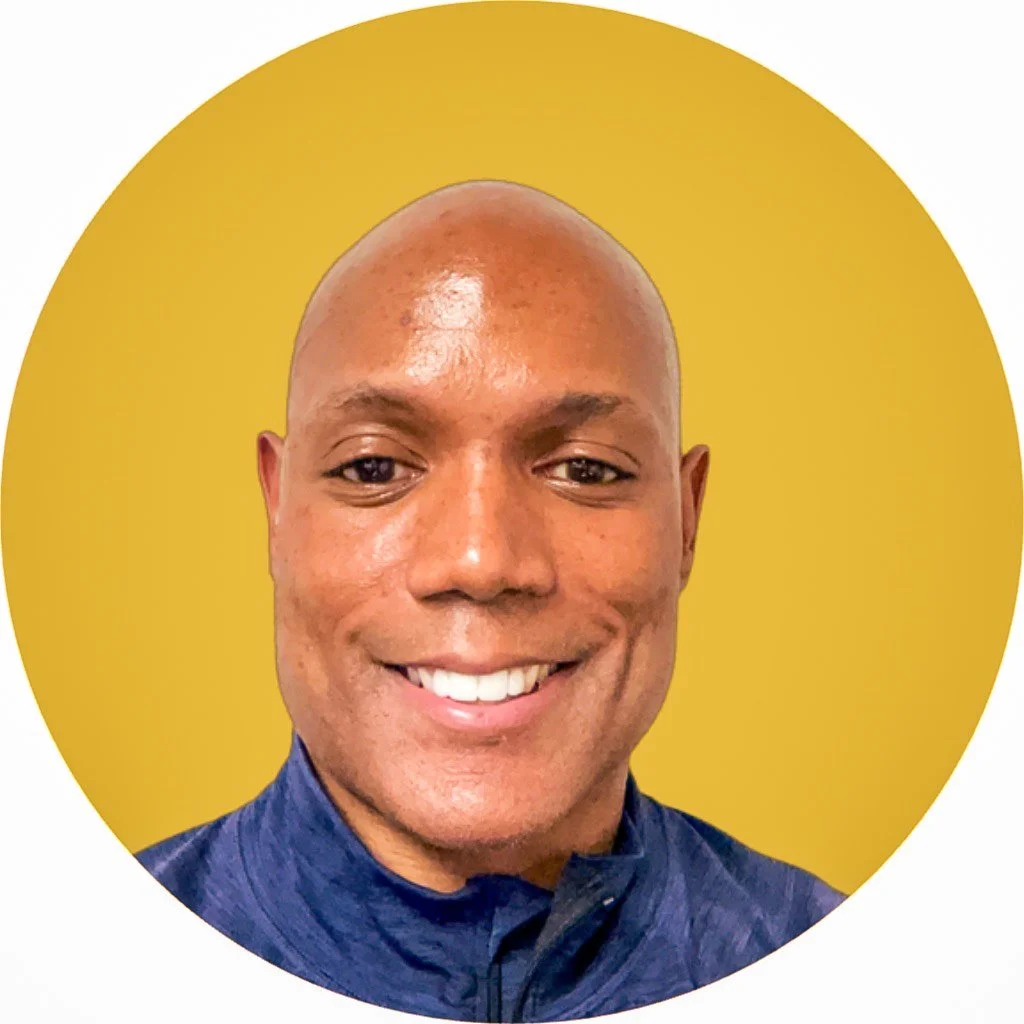 A smiling man with a bald head, dark eyes, and dark skin, wearing a dark blue shirt with a high collar, against a yellow circular background.