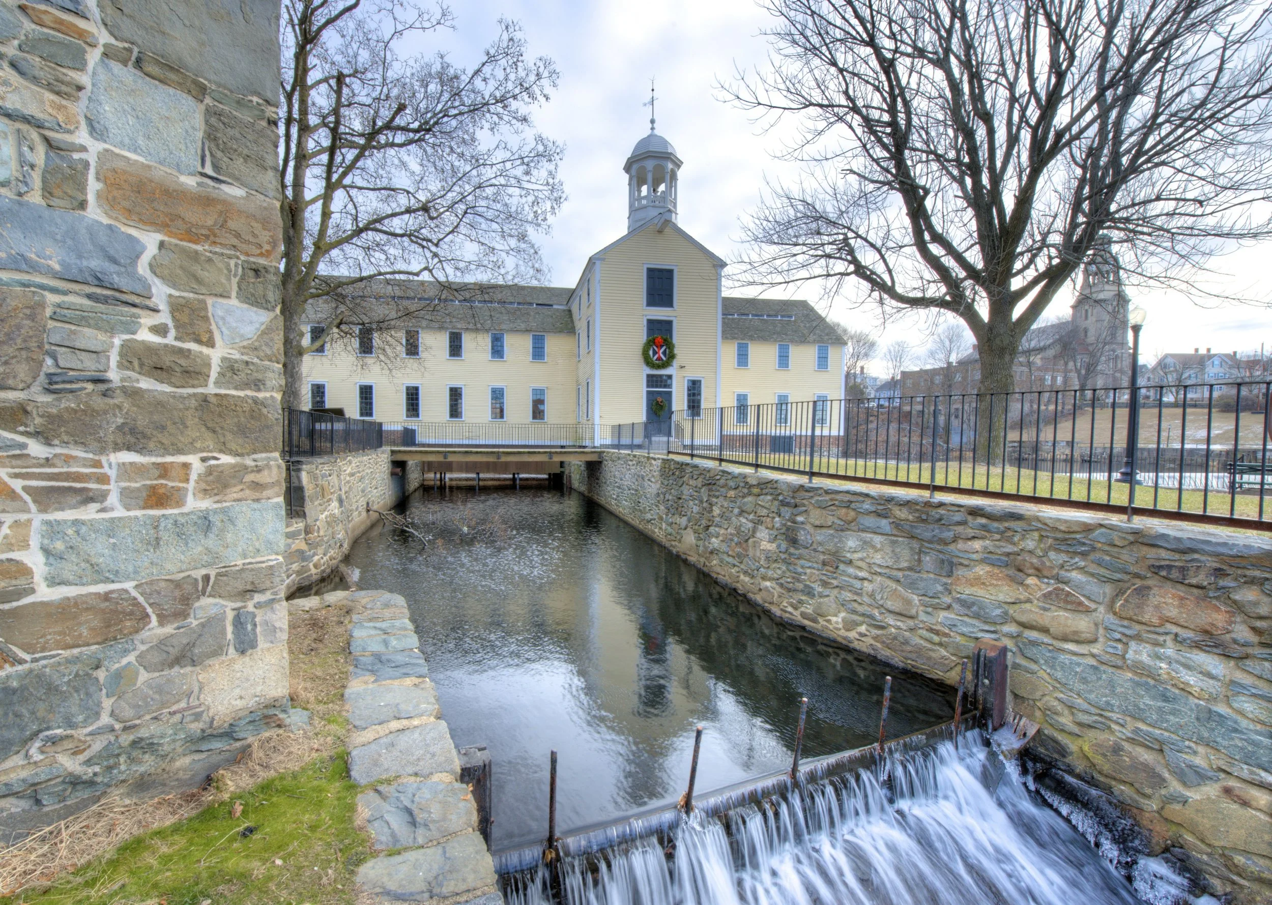 A small overpass with a stone wall on each side crosses over a narrow stream. On the far side of the stream, there's a large yellow building with blue window shutters, a clock tower, and holiday wreaths. Bare trees surround the scene, indicating late fall or winter. The sky is overcast.
