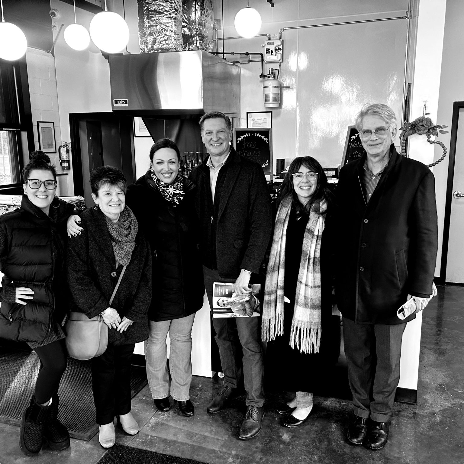 Group of six people standing together indoors, smiling, with a sign that reads 'The House' behind them.