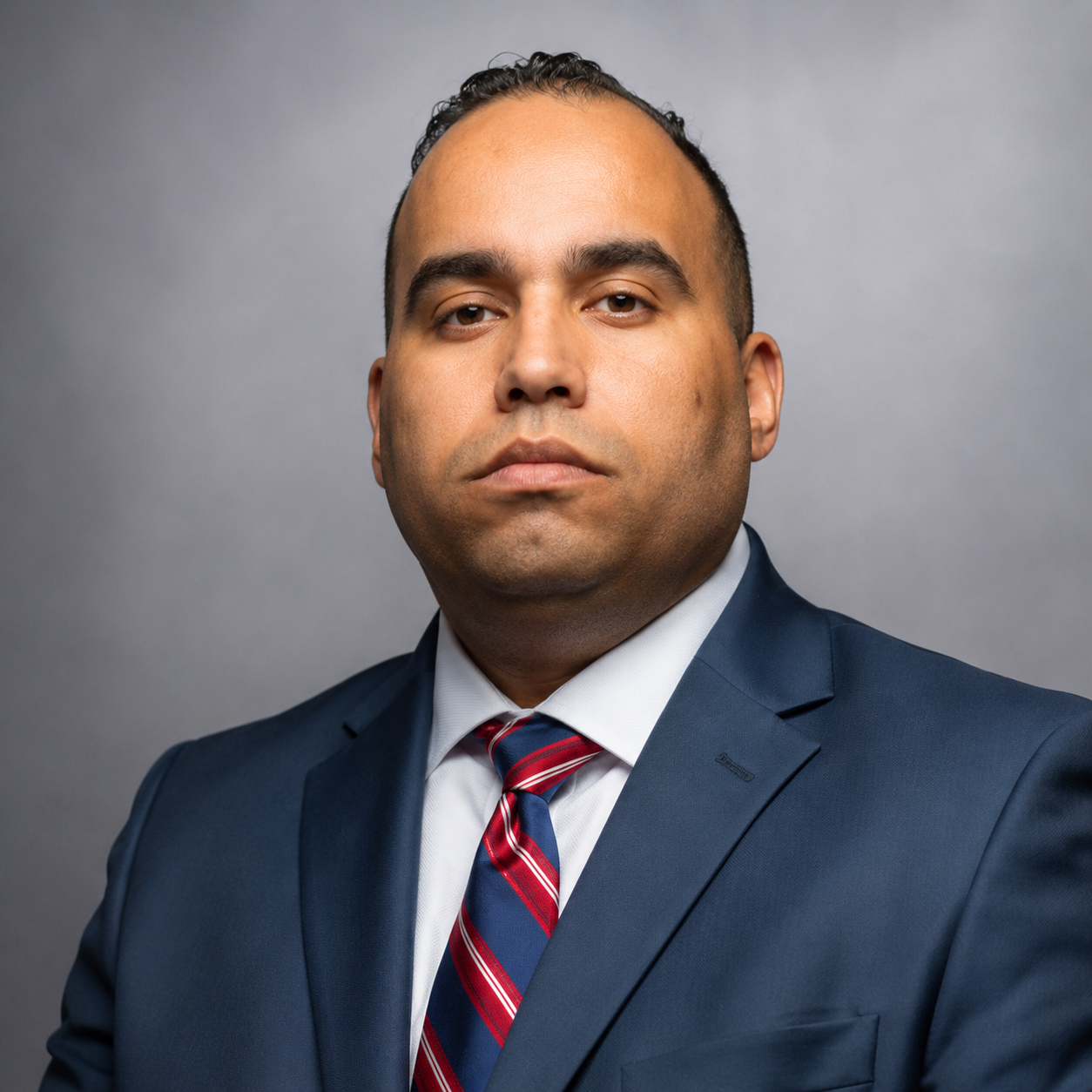 A man in a navy blue suit, white shirt, and red striped tie posing against a gray background.