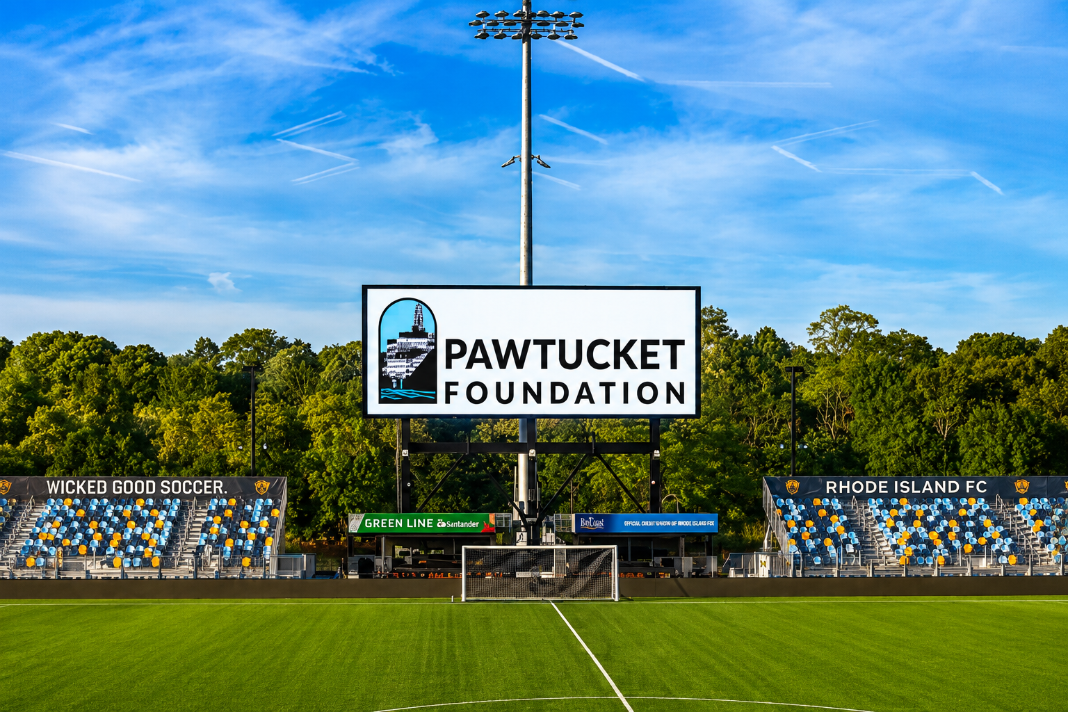 Empty soccer field with goal and stadium seating, featuring signs for Pawtucket Foundation, Wicked Good Soccer, and Rhode Island FC, under a clear blue sky.