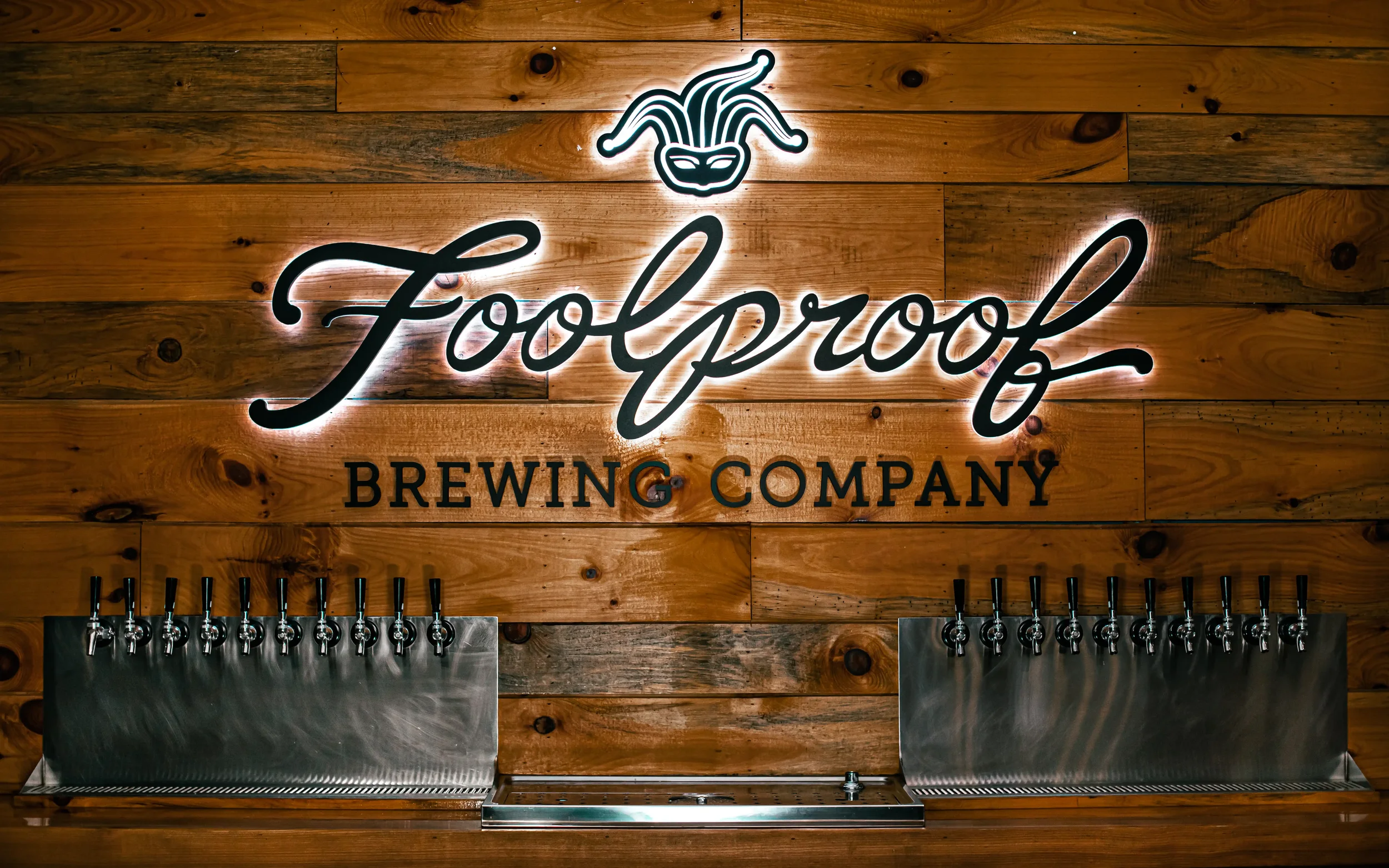 A wooden wall with a neon sign that reads 'Foolproof Brewing Company' and a stylized jester face logo above the text. Below the sign, there are several beer taps.