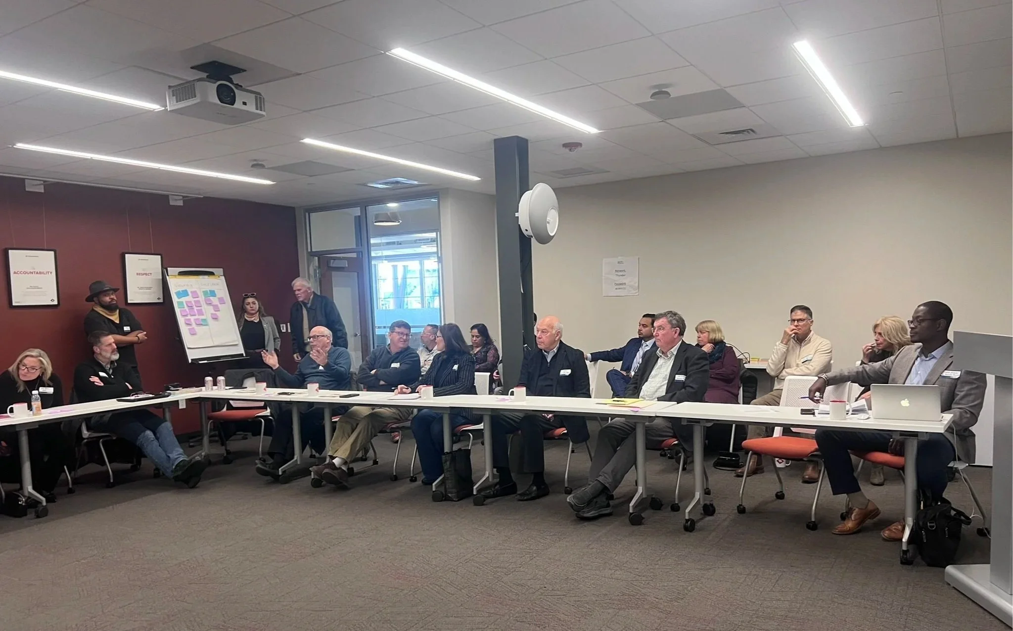 People attending a business meeting or conference in a modern conference room, seated around a long table, some taking notes, with a presentation or discussion occurring.